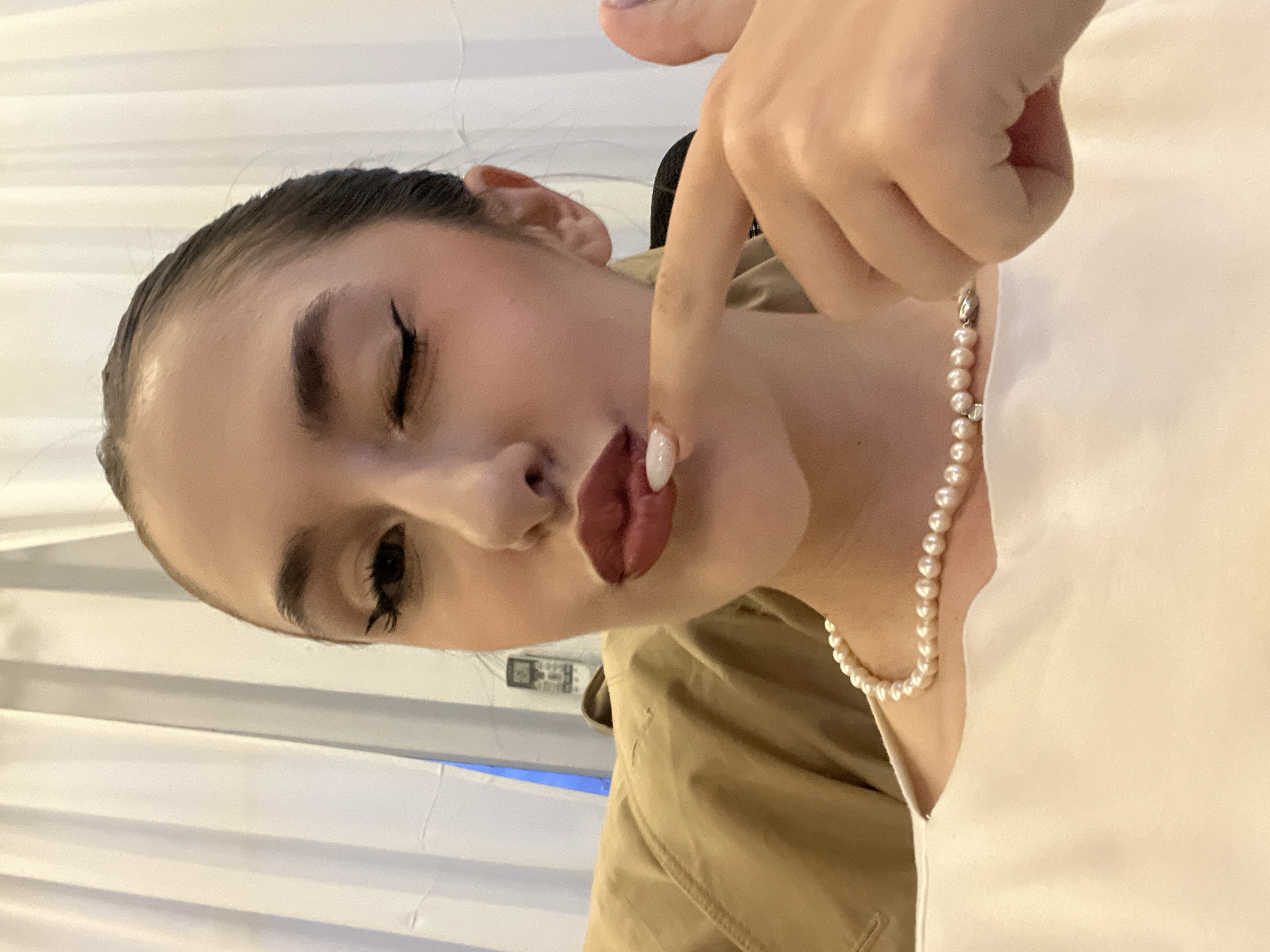 A close-up, portrait-oriented shot captures a young woman from the chest up, with a playful and suggestive expression. Her head is tilted slightly. She has dark hair pulled back, showcasing her face. Her makeup includes prominent winged eyeliner on both eyes, with one eye distinctly winked shut, and the other open and looking directly towards the viewer. Her lips are full, adorned with dark red lipstick, and her right index finger is gently pressed against her lower lip. The fingernail on this finger has a white polish.
Around her neck, she wears a delicate pearl necklace, consisting of small, uniformly round, white pearls. She is dressed in a light-colored top, possibly white or cream, and a segment of a beige or light brown jacket is visible on her left side.
The background is predominantly white, featuring subtle vertical lines or folds, suggesting an indoor setting with a wall or blinds. An indistinct, rectangular object, possibly a control panel or switch, can be faintly seen in the distant background over her left shoulder. The lighting is soft and even, highlighting her facial features. There is no visible text in the image. The general atmosphere conveyed is one of casual intimacy or flirtation. While the location context is Sofia, Bulgaria, there are no specific visual cues in the image to confirm this.