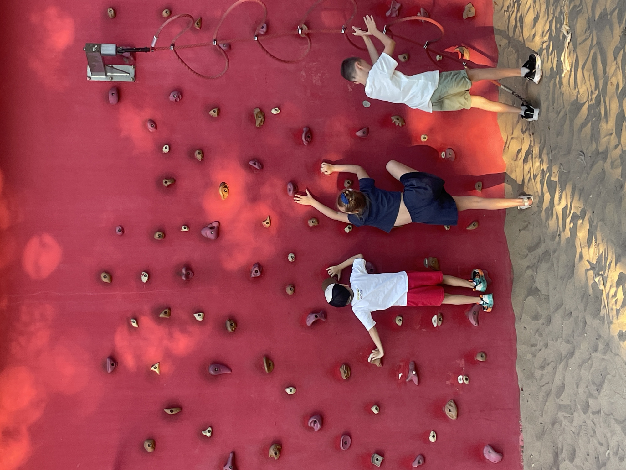 The image shows three children climbing a red climbing wall. The climbing wall has many different colored hand and foot holds. There are ropes attached to the wall. The ground in front of the wall is covered in sand. The children are wearing casual clothing. The lighting suggests it is daytime, possibly late afternoon due to the shadows. The image does not provide any information about the location in the city of Krasnodar, Russia.
