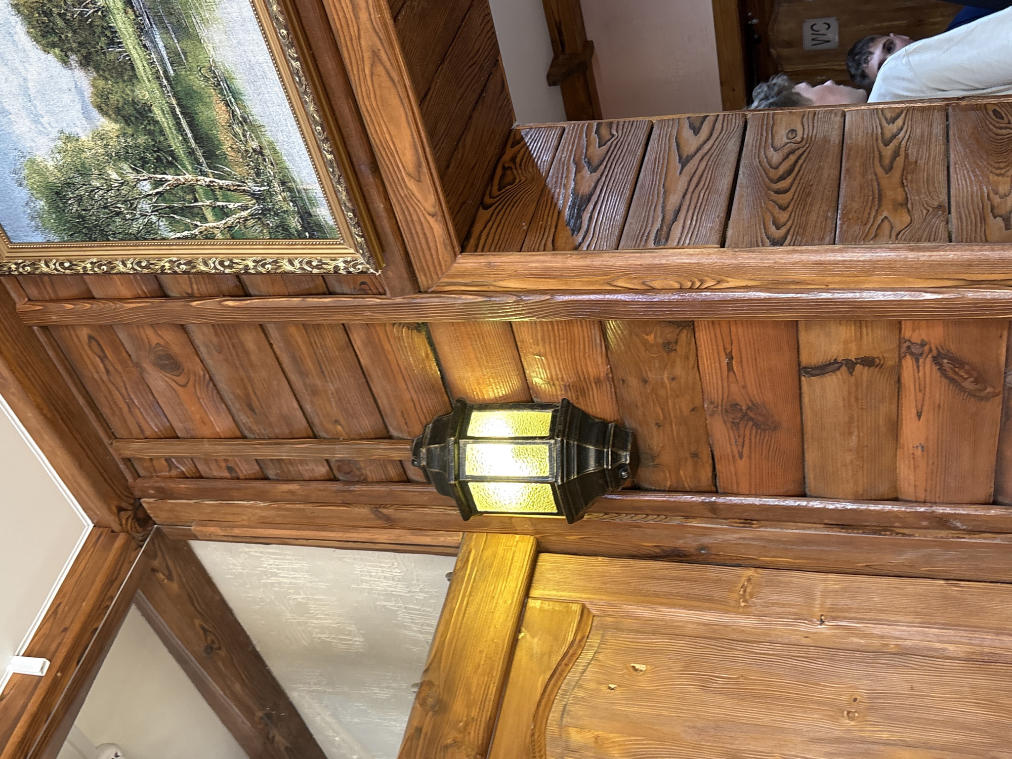 The image shows an indoor setting with a wooden ceiling and walls. A framed landscape painting hangs from the ceiling on the left side. A black, rectangular light fixture with yellow glass panels is mounted on the ceiling. Above the light fixture, a wooden railing or structure can be seen with people on the other side; one is wearing a white shirt. A sign labeled "WC" is visible above these people. The scene is likely in a building in Grishanki, Russia.