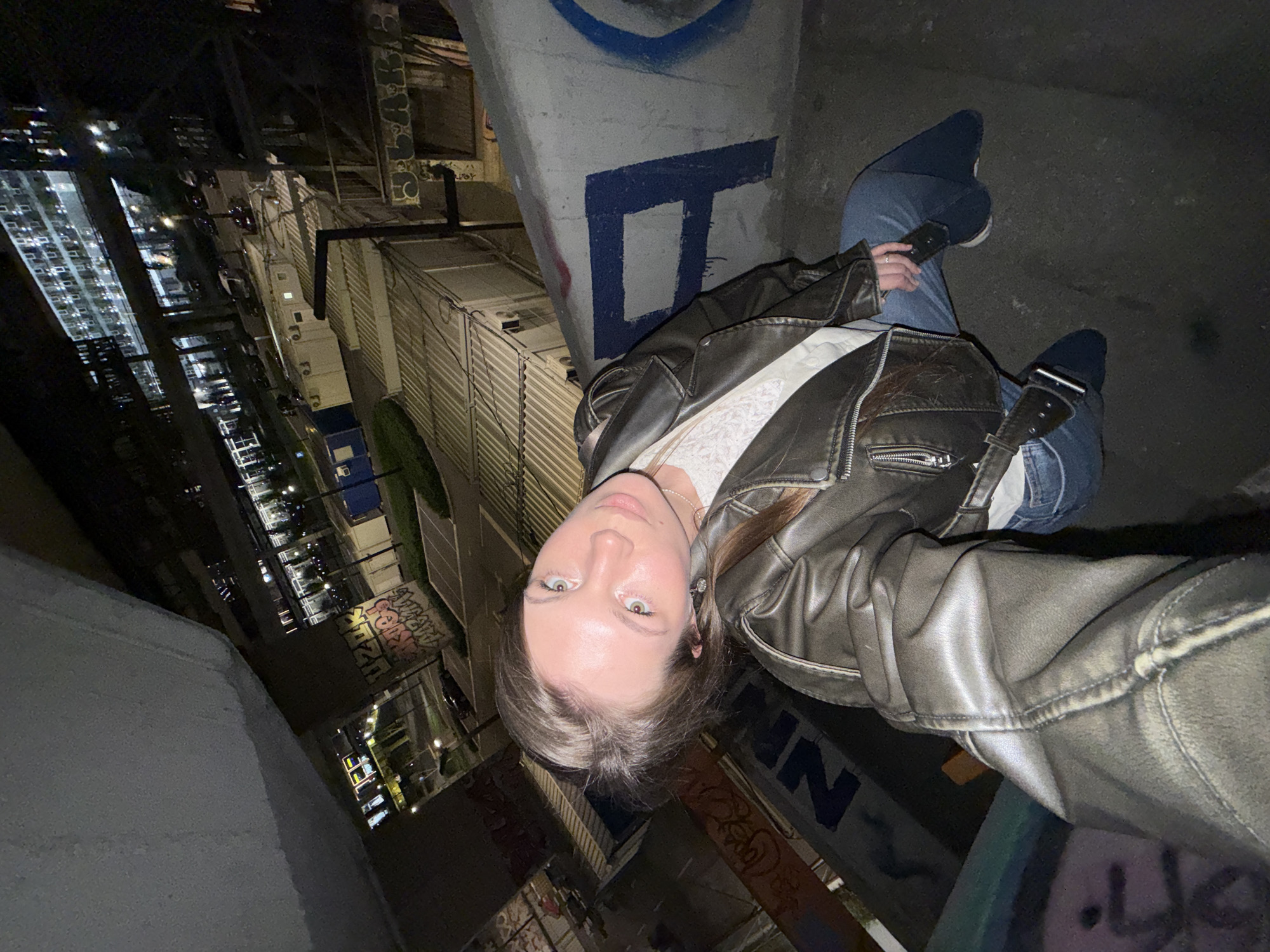 An overhead-oriented photograph, taken at night, captures a young woman in an urban setting. The image is actually a selfie taken by the woman, who appears to be leaning or sitting on a concrete structure with extensive graffiti.
The main subject is a woman with light-colored eyes and brownish hair, looking directly at the camera with a neutral expression. She is wearing a dark, possibly leather, jacket over a white top, blue jeans, and dark shoes. She holds a dark-colored smartphone in her left hand, which she is likely using to take the photo.
The setting is an elevated part of a city at night, possibly a rooftop or an overpass, in Belgrade, Serbia. The background features several tall buildings with illuminated windows, indicating a bustling cityscape. Below, there are visible streets with artificial lights and other urban structures, including what appear to be stacked industrial units or air conditioning components. The environment has a somewhat industrial and gritty feel, characterized by bare concrete walls and metal railings.
Notable details include the time of day, which is clearly night, evidenced by the darkness and the reliance on artificial light sources. The lighting creates strong contrasts and shadows throughout the scene. Numerous instances of graffiti are visible on the concrete surfaces. Prominently, a large blue letter, possibly a "D", is painted on the concrete pillar next to the woman. Other blue letters resembling "NN" are on a lower wall, and more colorful, illegible graffiti is seen further in the background, including words that might be "COLOR" and "KING". The woman's pose suggests a moment of rest or contemplation amidst the urban night.