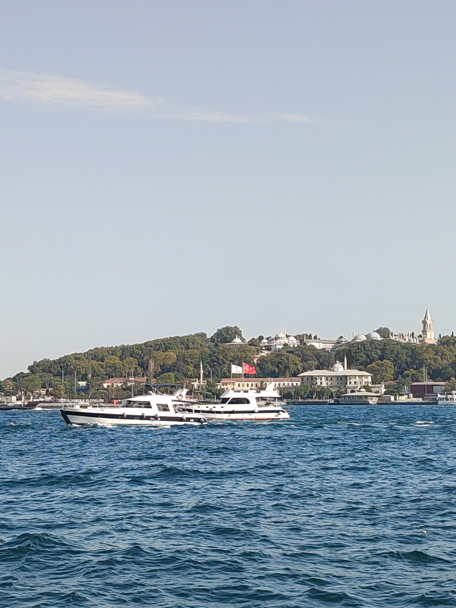 The image shows a body of water with two white boats in the foreground. In the background, a tree-covered hill rises, with buildings and domes visible on its crest. A minaret can be seen on the right side of the hill, and a Turkish flag flies on the left. The sky is clear and light blue. The scene depicts a coastal view in Beyoğlu, Turkey, likely the Bosphorus Strait, with Topkapi Palace and other historical structures in the background.
