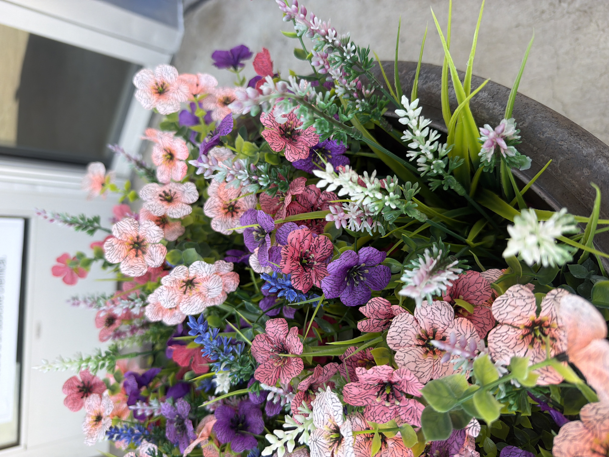 The image shows a close-up of a planter filled with artificial flowers in various shades of pink, purple, and blue, with green foliage. The planter itself is dark, weathered, and appears to be made of a textured material like concrete or resin. In the background, a portion of a white structure is visible, possibly a building or a door frame, with a dark reflective surface, suggesting a window or glass. The scene is well-lit, likely outdoors in daylight, based on the lighting and the reflective surface. There are no people or animals visible in the image. The overall impression is of a decorative element placed outdoors, likely near an entrance or walkway. The metadata indicates the image was taken in Lebanon, United States of America.