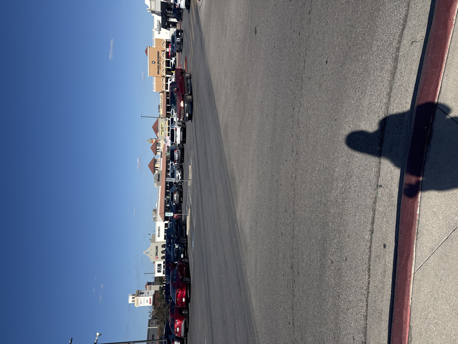 A street scene on a sunny day, featuring parked cars lining buildings with various retail signs. The sky is clear and blue. A wide asphalt road stretches into the distance, marked with faint lines. A red curb separates the road from a sidewalk where a person's shadow is cast.