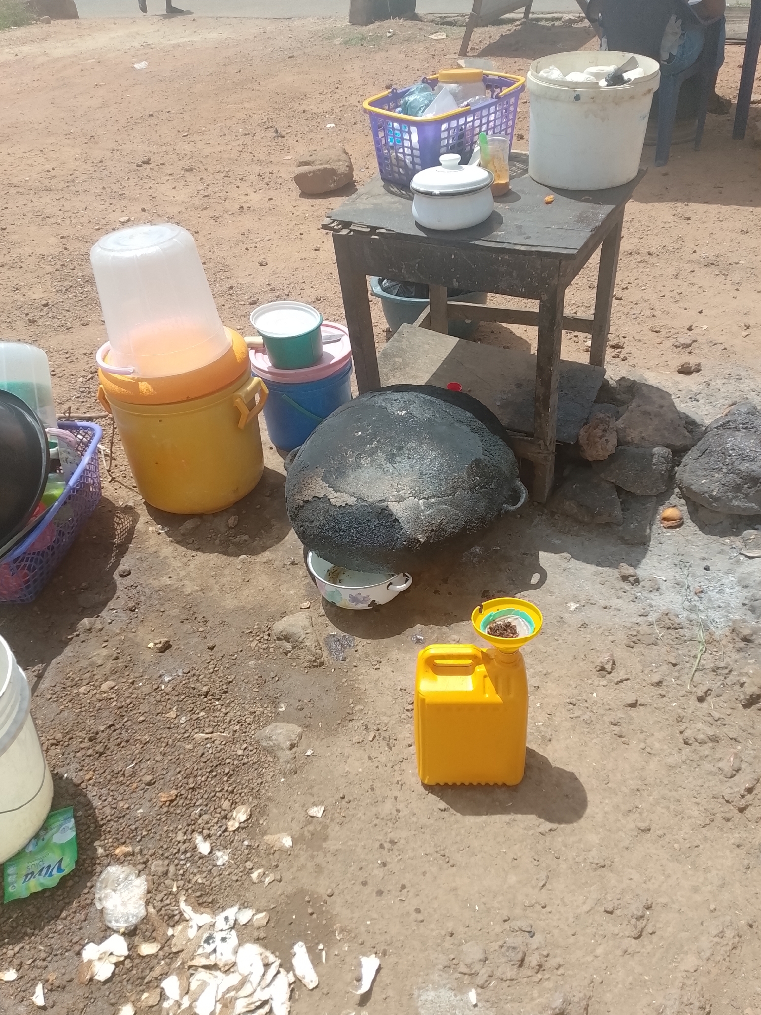 This image captures a vibrant outdoor cooking or vending setup on a dusty, unpaved ground, characteristic of a roadside stall in Jos, Nigeria. Bright sunlight illuminates the scene, indicating it is daytime.
In the foreground, a yellow plastic jerrycan stands on the dirt, topped with a yellow funnel containing dark brown granules, possibly spices or coffee beans. The ground around it is covered in loose soil, small stones, and scattered debris, including what appears to be discarded food peels and plastic waste. A white bucket, partially visible on the lower left, bears the legible text "VIVA plus," suggesting it once contained a detergent product.
The focal point in the mid-ground is a large, dark, heavily sooted cooking pot, likely cast iron, resting precariously on a makeshift stove formed by large rocks, indicating an open-fire cooking method. A small, patterned bowl sits directly beneath it. To the left, a large yellow insulated dispenser with a translucent white top holds a light orange liquid, likely a beverage. Beside it are smaller, colorful plastic buckets in green, pink, and blue. A purple plastic basket filled with various kitchen items, including pans, sits on the ground.
To the right, a sturdy but aged wooden table holds additional items for food preparation or serving. On the table, a white lidded pot, a clear plastic container holding an orange liquid, and a larger white bucket filled with cloths or other supplies are visible. Another purple basket sits on the table. In the background, a person's legs are partially visible walking on the dirt road, and a blue plastic chair can be seen behind the table. The scene portrays a functional, busy street food or beverage vendor's operation.