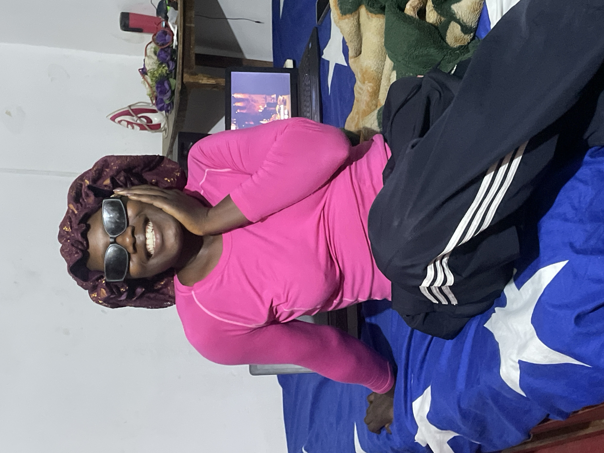 The image shows a young Black person lying down on a bed, wearing a pink long-sleeved shirt, dark pants with white stripes, and a dark, patterned bonnet. They are also wearing large sunglasses and smiling broadly, with their hand near their face. In the background, a laptop screen is visible, displaying an image of a cityscape with lights. There are also blankets and bedding on the bed, one with a blue background and white star patterns, and another with a green fuzzy texture. A wooden shelf is mounted on the wall above the bed, holding various items including a red and white object and what appears to be dried flowers or decorative elements. The scene suggests a casual, indoor setting, likely in a bedroom. The lighting is not strong, indicating it could be evening or a dimly lit room. The person's expression conveys happiness and a relaxed mood. The image appears to be taken in Jos, Nigeria, based on the provided location context.