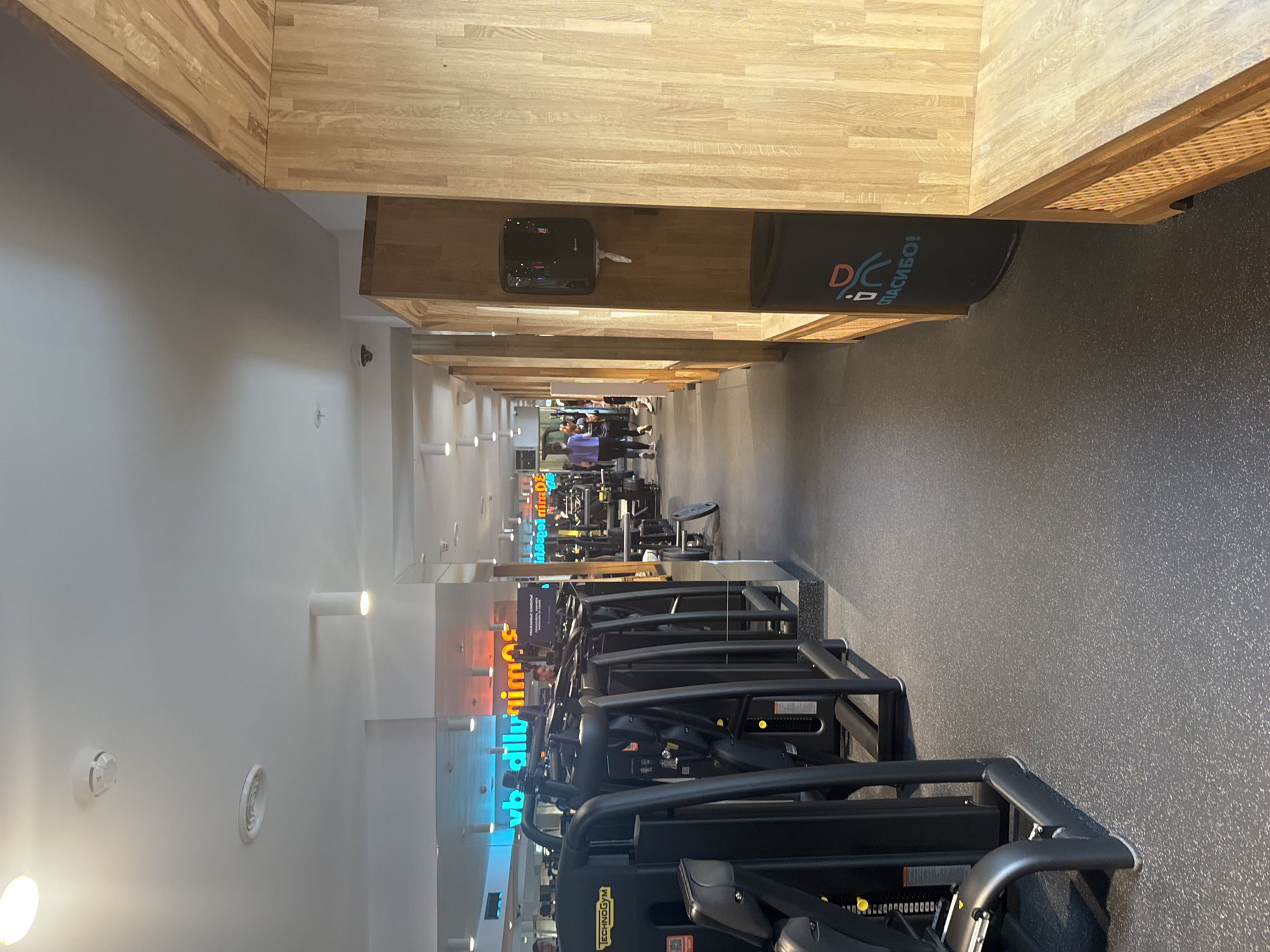 The image depicts the interior of a modern gym in Saint Petersburg, Russia. The scene is dominated by rows of exercise machines, predominantly black and manufactured by Technogym, stretching down a long, well-lit hallway. The gym features a clean, contemporary aesthetic with light-colored wooden paneling on the ceiling and upper walls, contrasting with the dark, speckled rubber flooring.
In the background, several people are visible, seemingly engaged in their workouts. One woman in a purple top and black leggings is walking, while others are blurred in the distance, suggesting movement and activity. The lighting is bright and even, indicating it is likely daytime or the gym is well-illuminated artificially.
Prominently displayed on a dark banner above a piece of equipment, there is text in Cyrillic: "СПАСИБО!". This translates to "Thank You!" and features a stylized logo with the letters "DC". On the machines themselves, "Technogym" is clearly visible. Other faint, colorful neon signs are also visible in the distance, though the text is illegible. The overall impression is one of a functional, active, and well-maintained fitness facility.
