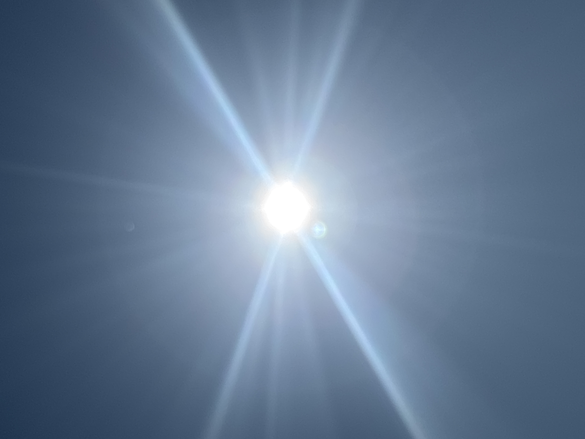 The image is a close-up, low-angle shot of the sun shining brightly in a clear blue sky. The sun is positioned in the center of the frame, appearing as a white, square-shaped object with strong rays of light emanating from it in all directions, creating a starburst effect. There are some lens flares visible, including a circular one slightly below and to the left of the sun, and a faint, almost imperceptible object, possibly the moon or a distant celestial body, appears as a small, pale circle in the upper portion of the sky. The sky is a uniform gradient of blue, lighter near the sun and darkening towards the edges of the frame. The overall impression is one of intense sunlight and a clear day. No specific landmarks or indications of the city of Arkhipovskoe, Russia are visible in this image.
