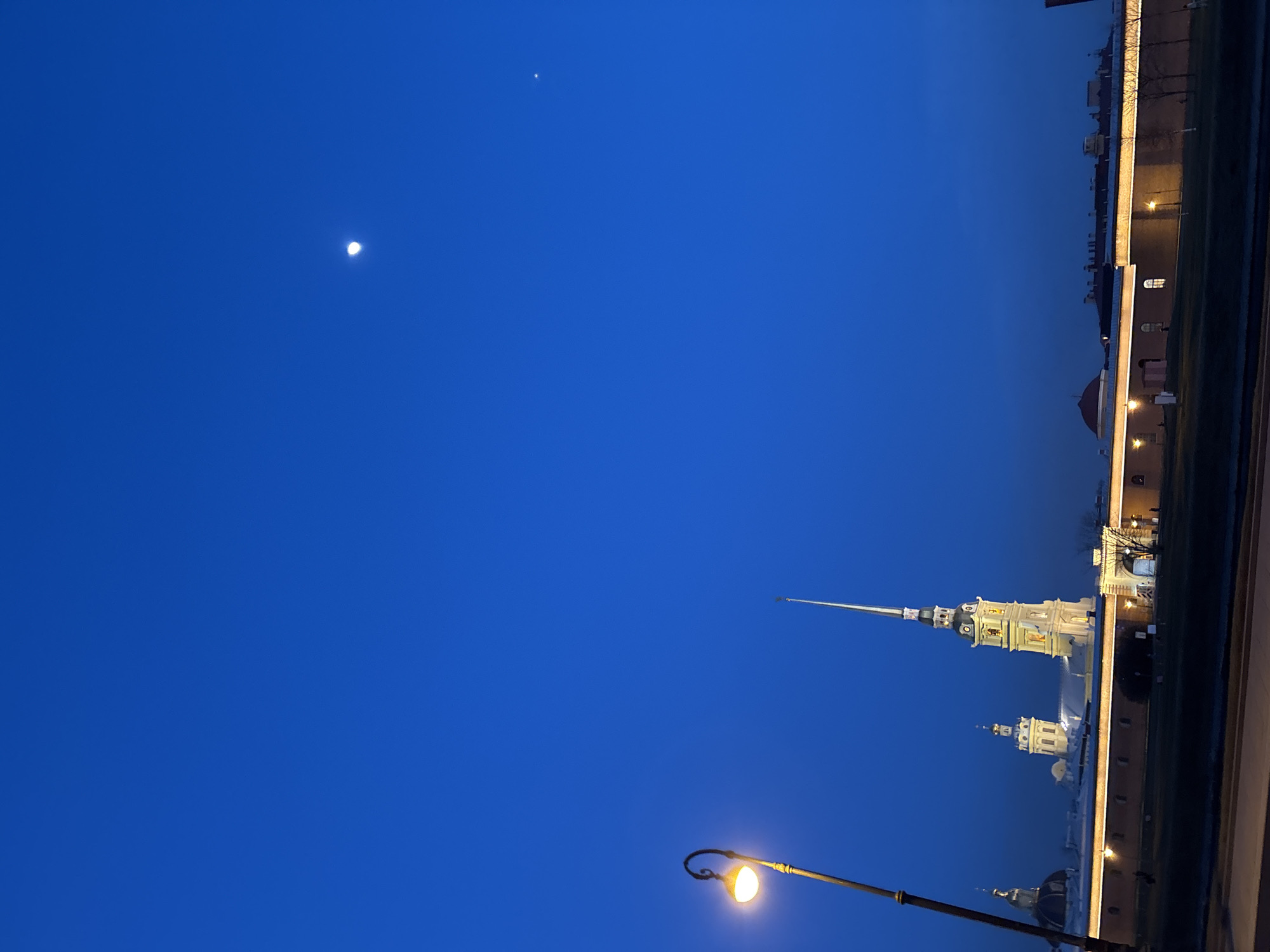 The image shows the Peter and Paul Fortress in Saint Petersburg, Russia, at twilight. The fortress's illuminated spire dominates the right side of the frame. The sky is a deep blue, with the moon visible in the upper left. A street lamp is lit in the lower right. In the background, the fortress wall stretches into the distance, illuminated by artificial lights.
