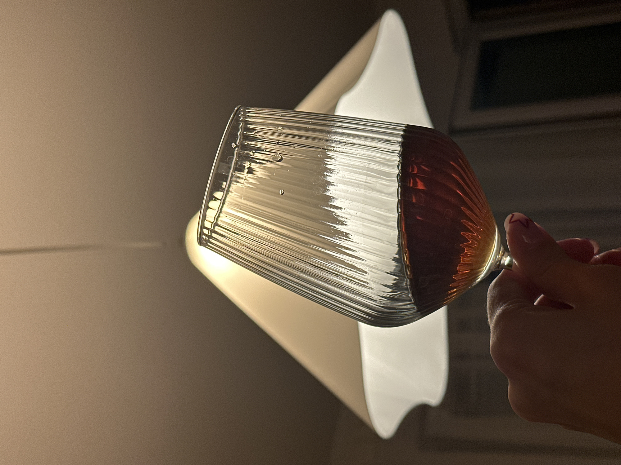 The image depicts a hand holding a ribbed glass filled with an amber-colored liquid. Behind the glass is a white, lamp shade. The background consists of neutral-toned walls and a window. There is no identifiable information suggesting that the image was taken in Novi Sad, Serbia.
