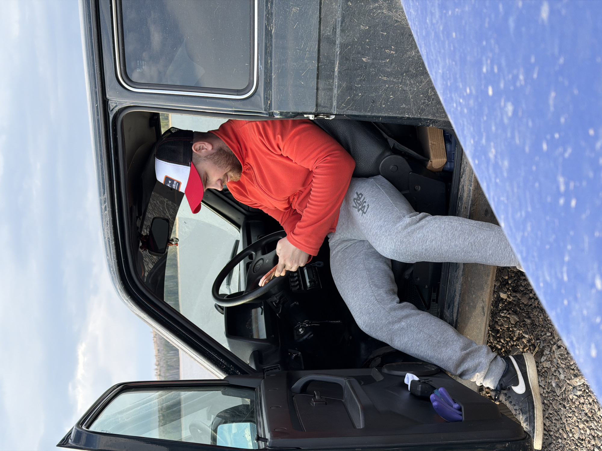 A medium-shot photograph depicts a man sitting in the driver's seat of a black vehicle, possibly a UAZ, with an open door. The man is wearing a red hooded sweatshirt, grey sweatpants, and black and white Nike shoes. He is also wearing a baseball cap with a red brim, white front panel, and black mesh back. The man is holding an object, possibly a pair of jumper cables, and his hand is on the steering wheel. The vehicle's door is open, revealing the outside environment, which appears to be a gravelly area near a body of water. The sky is overcast. The location is possibly in the city of Chaikovsky, Russia. The photograph is slightly rotated counterclockwise.
