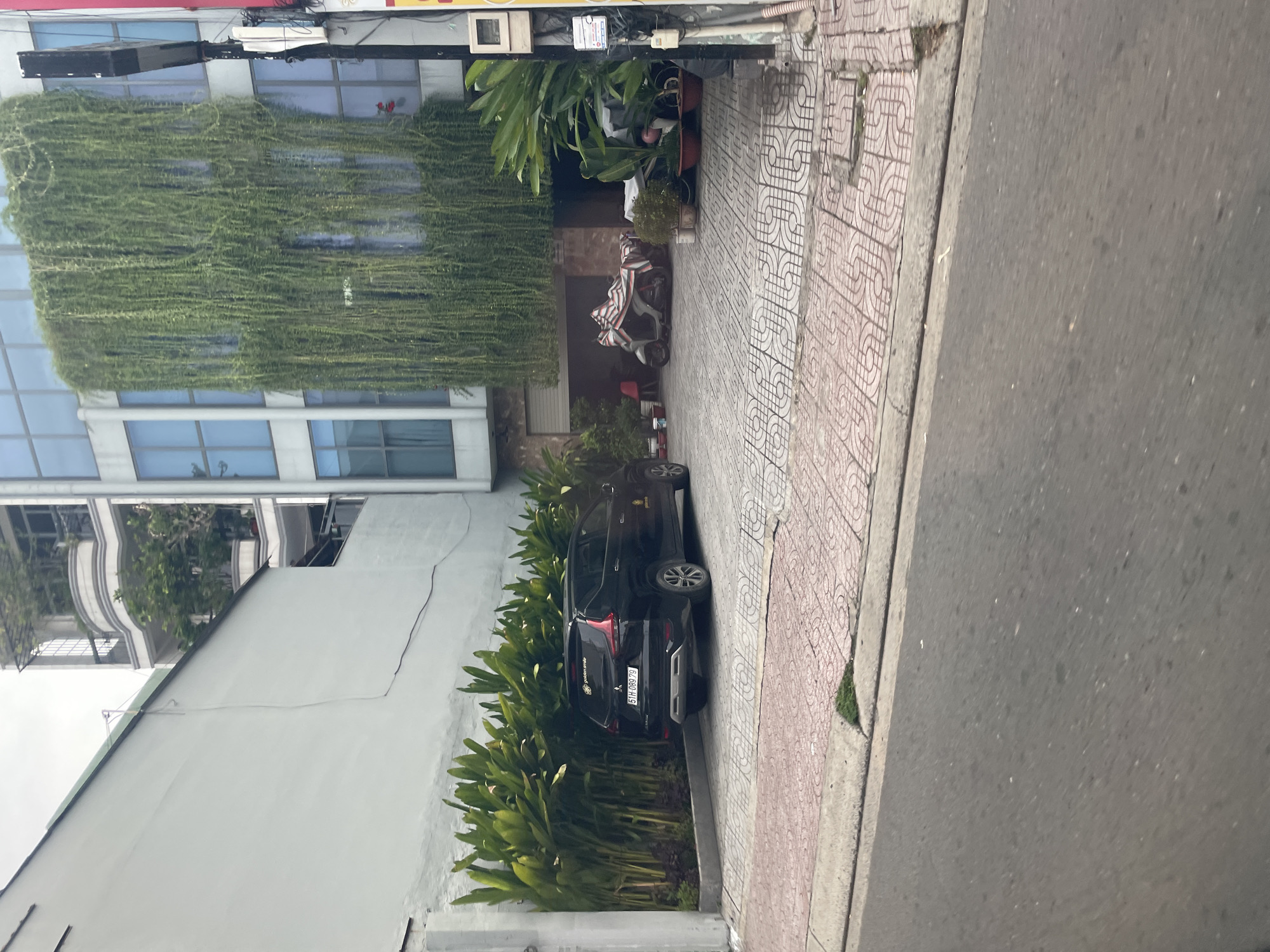 The image shows a street scene in Thủ Đức, Vietnam. In the foreground, a paved road transitions to a sidewalk with a decorative tiled pattern. Beyond the sidewalk, a parked black SUV is positioned in a driveway. To the left of the SUV, a dense row of tall green plants lines a white wall. To the right of the driveway, a building with a modern facade covered in hanging green vines and large blue-tinted windows is visible. Partially obscured by the plants and driveway, a scooter is parked under a covered area in front of the building. The overall environment suggests an urban setting with a mix of residential and commercial elements.