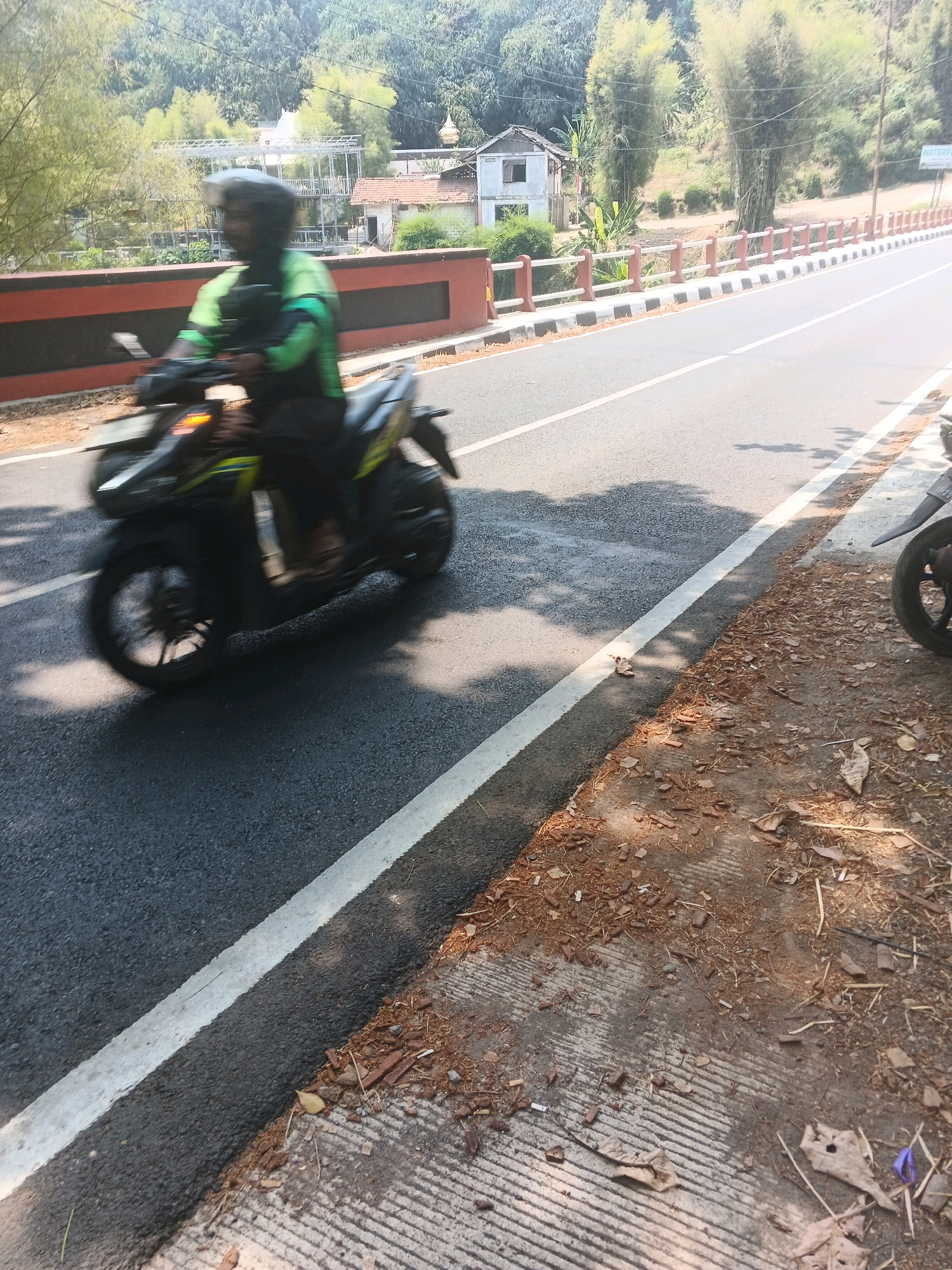 A blurry, motion-filled photograph captures a motorcycle rider on a paved road in a tropical environment. The rider, wearing a helmet and a green and black jacket, is on a dark-colored scooter. The scooter's headlight is illuminated. The road has a white dividing line and a concrete shoulder littered with dry leaves and debris. In the background, a red and black barrier runs alongside the road, separating it from a sloped embankment covered in lush green vegetation and trees. A few simple structures, including a house with a red tiled roof and a metal frame building, are visible amidst the greenery. The overall scene suggests a sunny day in a semi-rural or suburban area, possibly near a bridge or overpass, consistent with locations in Semarang, Indonesia.