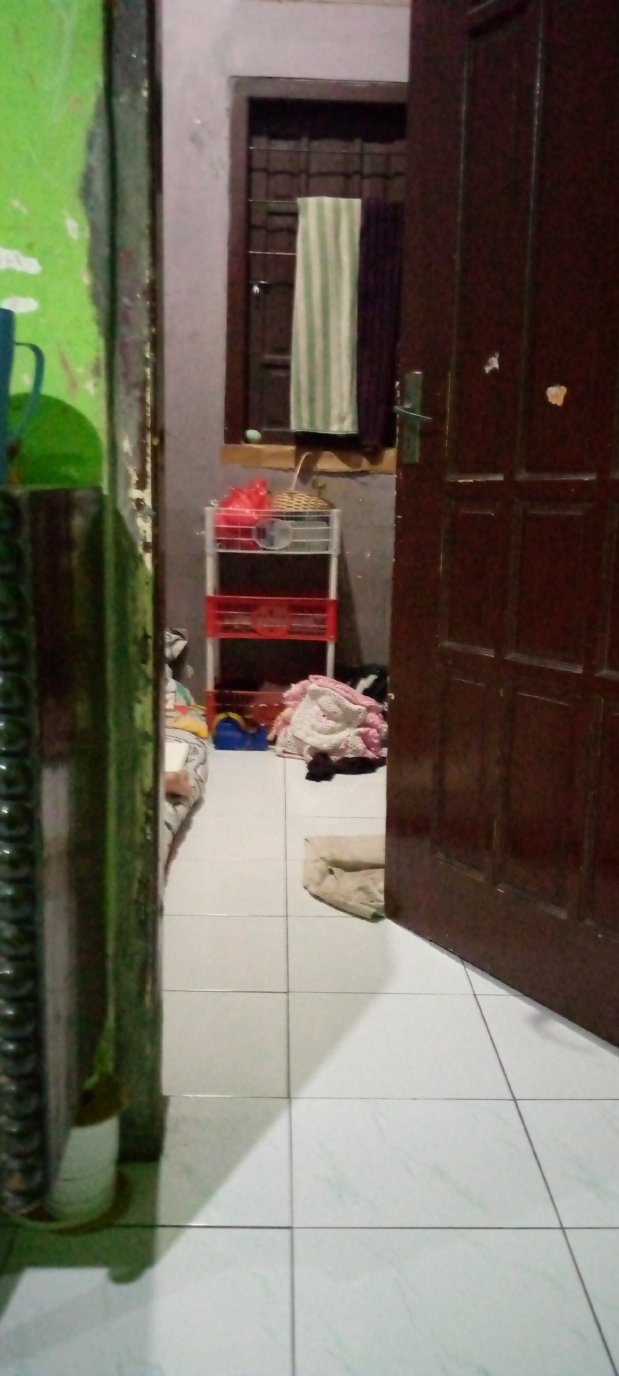 The image depicts an interior scene, possibly a hallway or entryway, in a residential setting. The floor is tiled with white and light green square tiles. To the left, a portion of a bright green wall is visible, alongside a dark, textured vertical element that appears to be a doorframe or pillar, with a potted plant at its base. In the center, a white multi-tiered shelf unit holds various items, including red plastic bags and a woven basket. A closed, dark brown wooden door occupies the right side of the frame, partially open, revealing a sliver of the interior. In the background, a window with a dark frame and barred panes is visible, with two towels, one striped green and white and the other purple, hanging in front of it. A pile of laundry, primarily pink and white fabric, is situated on the floor near the shelf unit, alongside a blue plastic object. A person's leg, clad in striped fabric, is partially visible on the floor to the left of the laundry pile. A light-colored folded cloth or blanket lies on the floor in the foreground. The overall impression is of a somewhat cluttered living space. No specific landmarks or context are provided to pinpoint the exact location within Sumberwono, Indonesia, beyond it being an indoor residential environment.