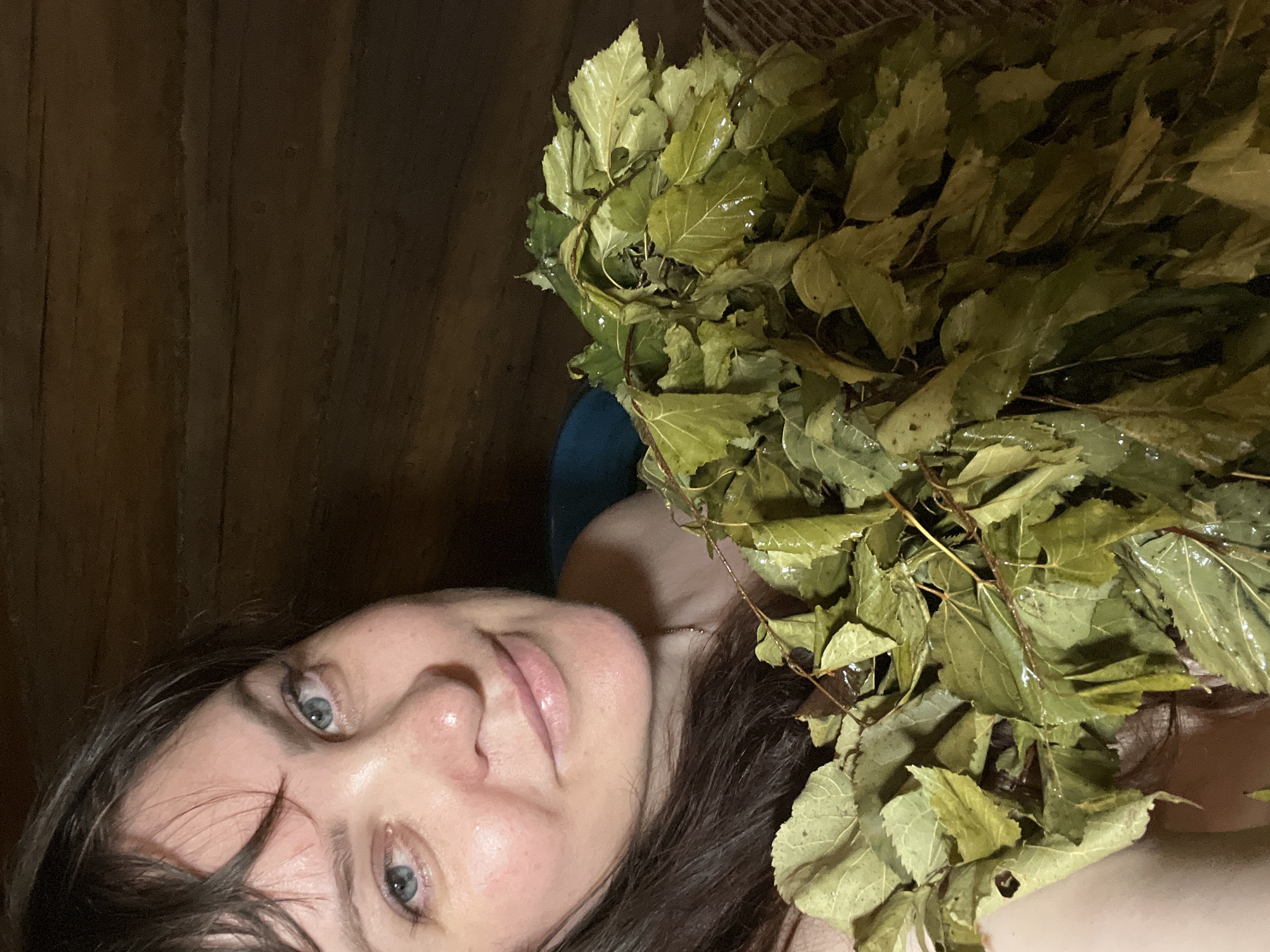 A woman with dark hair and blue eyes, smiling and holding a bunch of wet green leaves in a wooden sauna. The woman is facing the camera and her face is partially obscured by the leaves. The background is composed of dark wooden walls. The image appears to have been taken in the city of Panteleevka, Russia.
