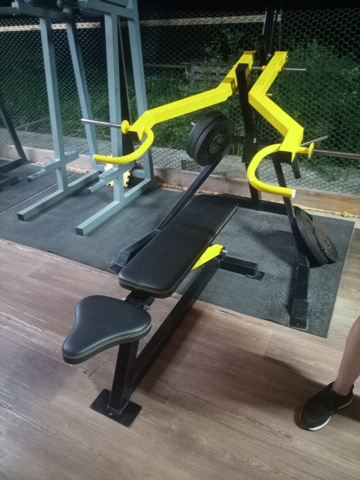 This is a medium-angle, full-shot photograph of a weightlifting bench with a weight rack. The weight rack is yellow and black, and the bench is black and yellow. The bench has a black padded seat and backrest, and a black metal frame. The weight rack has yellow arms and a black frame. There are black weight plates on the rack. The scene is indoors, with a wooden floor and a black rubber mat under the equipment. A person's legs and feet, wearing black sneakers, are visible in the lower right corner of the image. The background shows a wire fence and greenery. The location is identified as a gym in Nalchik, Russia.