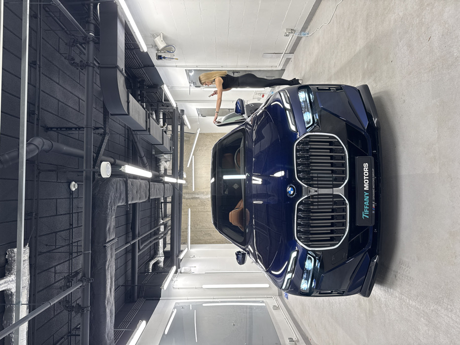 A blue BMW car is parked in a modern, well-lit garage with a woman in a black outfit leaning on the car door while looking at her phone. The garage has a gray concrete floor and white walls illuminated by fluorescent lights. Black pipes and ducts run along the ceiling on one side of the car. "TIFFANY MOTORS" is displayed on a logo affixed to the car's front grill.