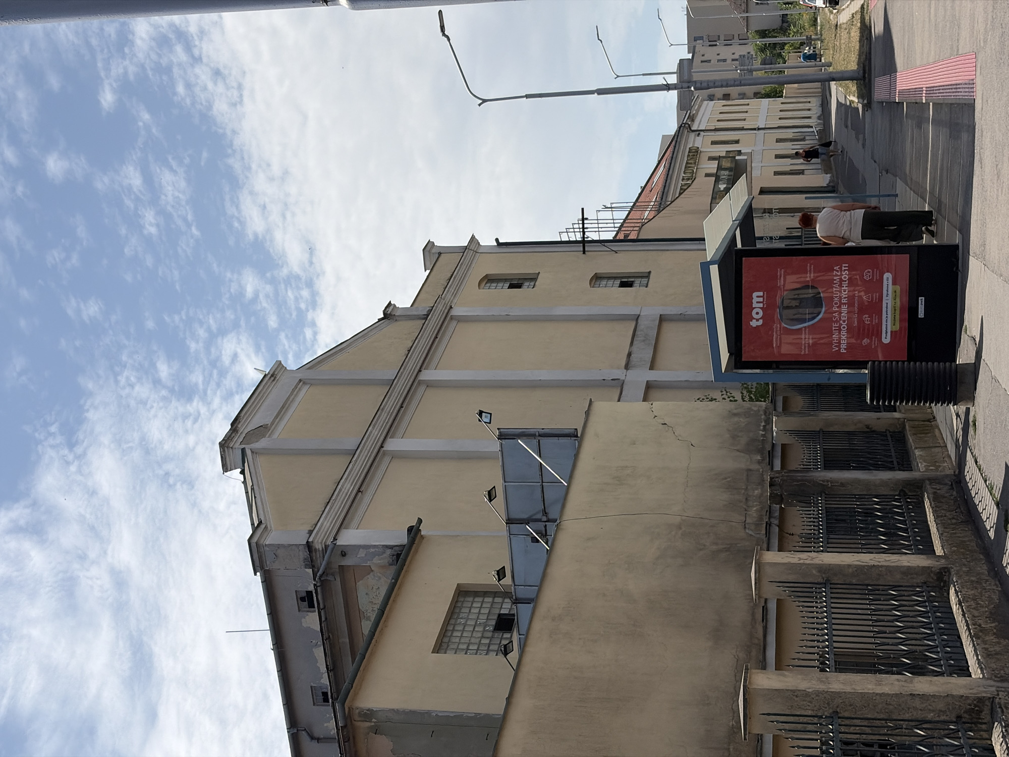 The image depicts a street scene in Nitra, Slovakia. A bus stop shelter with a red advertisement for "tom" is positioned on the right side of the frame. The advertisement features text in Slovak, including "VYHNITE SA POKUTAM ZA PREKROCENIE RYCHLOSTI," which translates to "Avoid fines for speeding." A woman wearing a light-colored shirt and dark pants walks away from the bus stop. In the background, there are several buildings, including a light-yellow building with a faded facade and a dark metal fence. The sky is blue with scattered white clouds. The street is paved, and there is a pink tactile paving area on the sidewalk.
