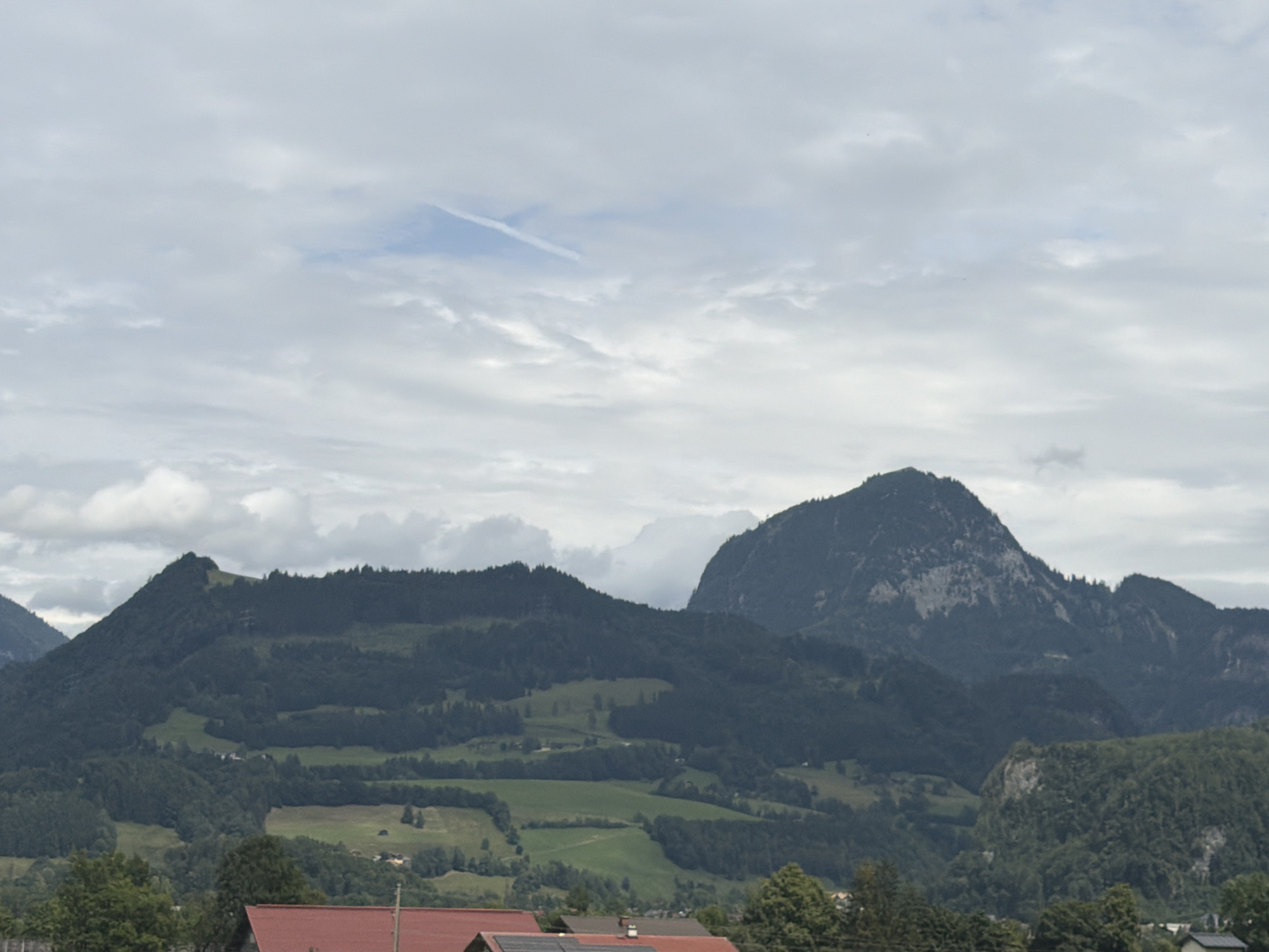 This media file captures a panoramic view of a mountainous landscape under a cloudy sky. The scene depicts several large, densely forested mountains that dominate the background. Patches of green fields and smaller trees are visible on the lower slopes of these mountains, suggesting a rural or alpine setting. In the foreground, the tops of buildings with red roofs are partially visible, hinting at human habitation. The sky is overcast with scattered clouds, with a faint contrail visible in the upper portion of the image. The overall impression is one of a serene, natural environment, possibly in the late afternoon or early morning due to the diffused light. The location context provided is Torren, Austria, which aligns with the mountainous terrain and architecture depicted. No people or specific activities are evident; the focus is on the grandeur of the natural scenery.