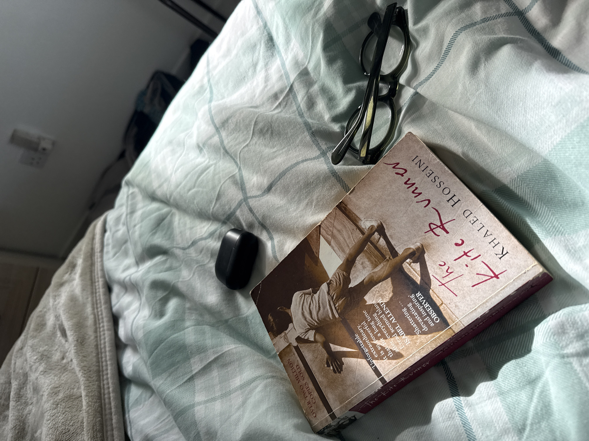 The image depicts a bed covered with a light blue and white plaid patterned sheet. On the sheet rests a copy of Khaled Hosseini's novel "The Kite Runner," a pair of black-rimmed eyeglasses, and a small black wireless earbud case. The book is prominently displayed, with its cover featuring an image of a person flying a kite. Part of a gray blanket is visible at the lower left of the frame. The environment appears to be indoors, likely a bedroom, given the bed and personal items.