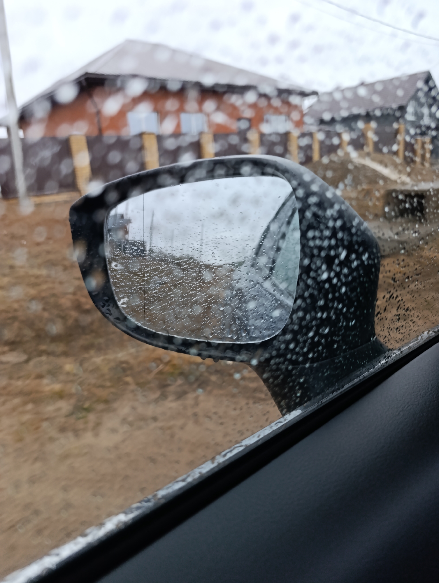 The image shows a car's side mirror covered in raindrops, reflecting a blurred view of a street in Chaikovsky, Russia. The mirror's frame is black, and the reflection shows a wet road and a house with a brown roof and light-colored siding. The background outside the mirror is also blurred due to raindrops on the window, but details of other buildings and street features can be made out. Inside the car, the door's interior panel is visible in the lower part of the image. The weather appears to be rainy.