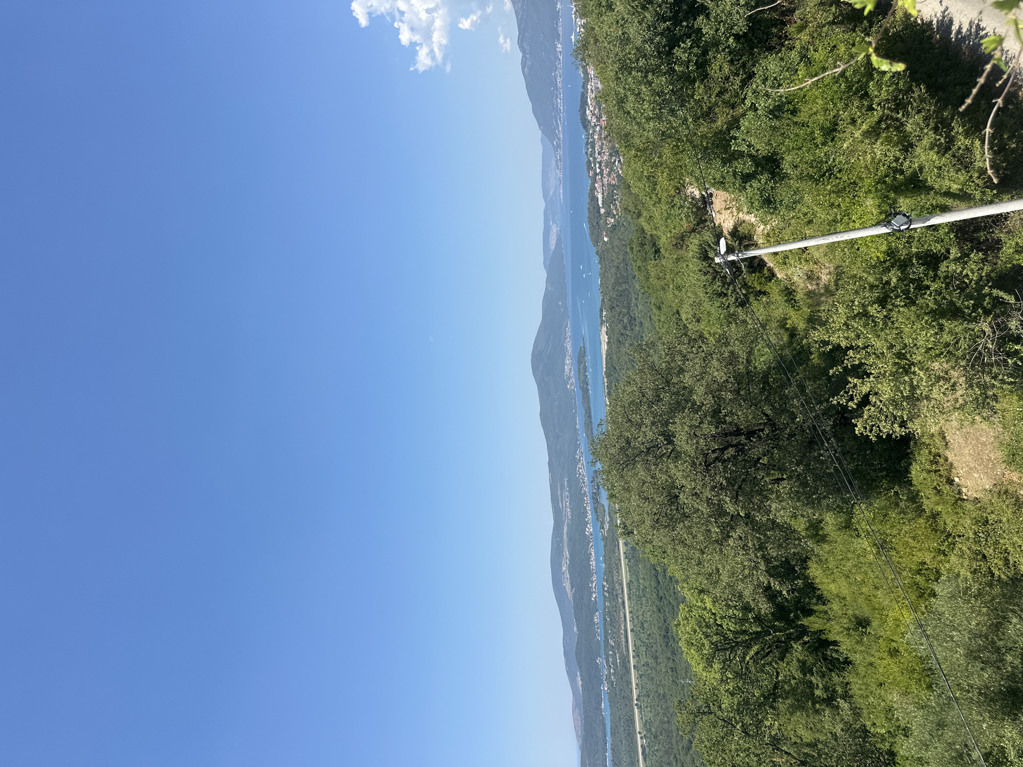 The image captures a panoramic view of a bay and mountainous landscape, likely from a high vantage point in Kavač, Montenegro. The sky is a clear, vibrant blue with a few wispy clouds on the right horizon. Below, a vast expanse of turquoise water stretches out, dotted with numerous boats of various sizes, suggesting active maritime activity. Along the shoreline of the bay, a town with red-tiled roofs and other buildings is visible, nestled against the backdrop of rolling green hills and distant, hazy mountains. The foreground is dominated by lush green foliage, dense trees, and shrubs, characteristic of a Mediterranean or Balkan environment. A single utility pole with wires runs diagonally across the lower right quadrant of the image, indicating the presence of infrastructure. The overall scene conveys a sense of natural beauty and a settled coastal community.