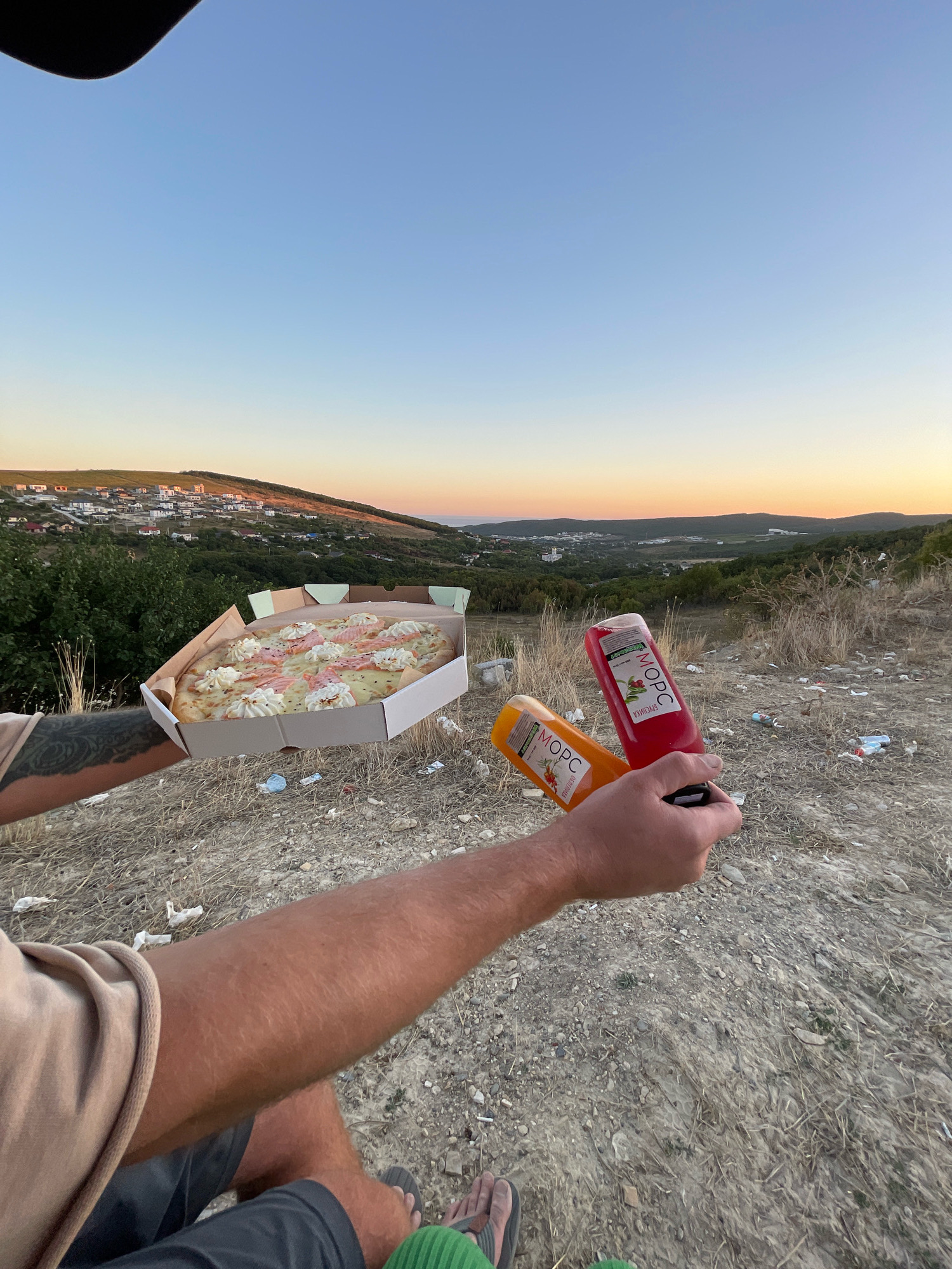 In Varvarovka, Russia, at dusk, a person holds a pizza and two bottles of "МОРС" juice overlooking a semi-rural town nestled in rolling hills. The pizza, in a white cardboard box, appears to be topped with cream, prosciutto, and possibly cheese. The two bottles of "МОРС" are one red and one orange. The ground is covered in dry grass, dirt, and scattered trash, including cigarette butts and plastic wrappers. In the distance, houses with red roofs dot the hillside, leading down to the coastline. The sky transitions from a clear blue at the top to a soft orange and yellow at the horizon.