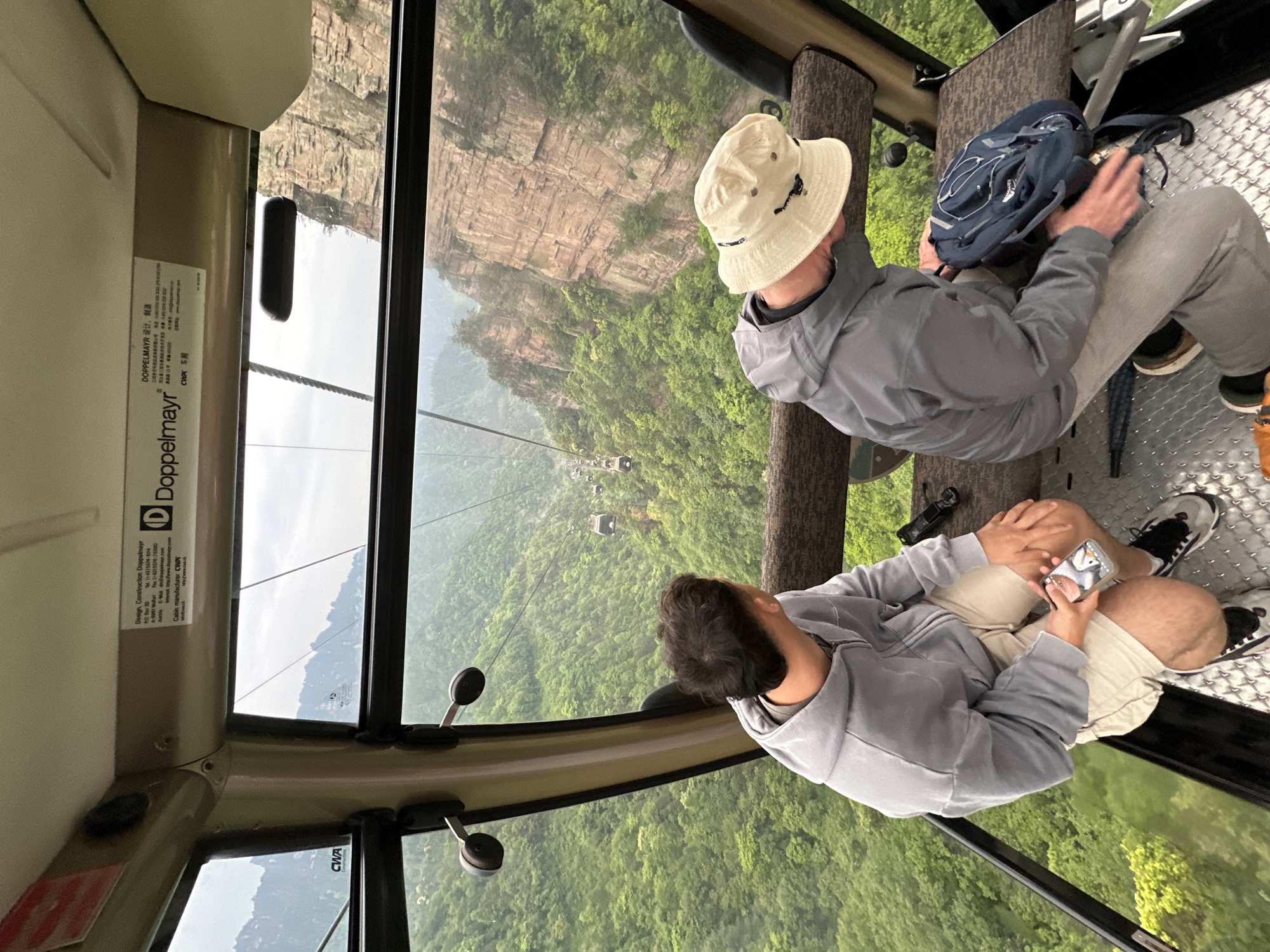 Captured from inside a cable car, the image presents an elevated view of Wulingyuan, China, characterized by towering sandstone pillars and lush green forests. Two individuals are seated in the cable car, one wearing a tan bucket hat and a gray jacket, while the other is in a similar gray jacket, holding a mobile phone. A blue backpack rests beside the first person, and a small black camera is seen near the second. The interior of the cable car is visible, including the metal floor and part of the framework. Outside the window, the landscape unfolds dramatically, with a series of mountains receding into the distance. The cable car system, with its suspension cables and distant cars, is also visible.
