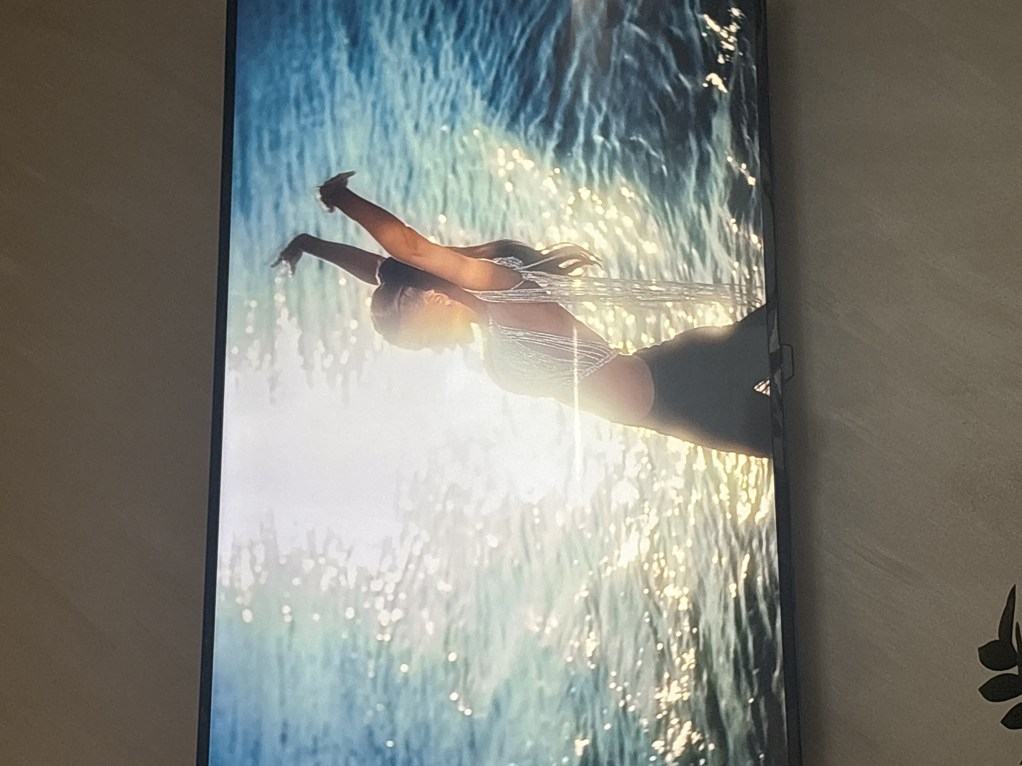 The media file shows a close-up, vertically oriented image of a television screen displaying a stylized image. The image on the screen features a person, likely a woman, in a graceful pose against a backdrop of shimmering water and sunlight. Her body is angled, with arms extended upwards and outwards, suggesting movement or a moment of suspended animation. She appears to be wearing a light-colored, possibly lacy or textured top and dark bottoms.
The background is dominated by what appears to be ocean waves or a large body of water, with the sunlight reflecting off the surface, creating numerous bright, sparkling highlights. The water transitions from darker blues to lighter, almost white areas where the light is most intense. A soft, golden glow emanates from the lower right portion of the image, suggesting it might be sunrise or sunset. There are no discernible text elements within the image on the television screen. The overall mood conveyed by the image is one of serenity, beauty, and perhaps athleticism or artistic expression. The television itself is a dark, rectangular screen set against a textured, neutral-colored wall, likely in an indoor setting. A small, dark object, possibly a plant with dark leaves, is partially visible in the lower right corner of the frame, outside of the television screen. The image appears to be a still from a video or a photograph displayed on the screen.