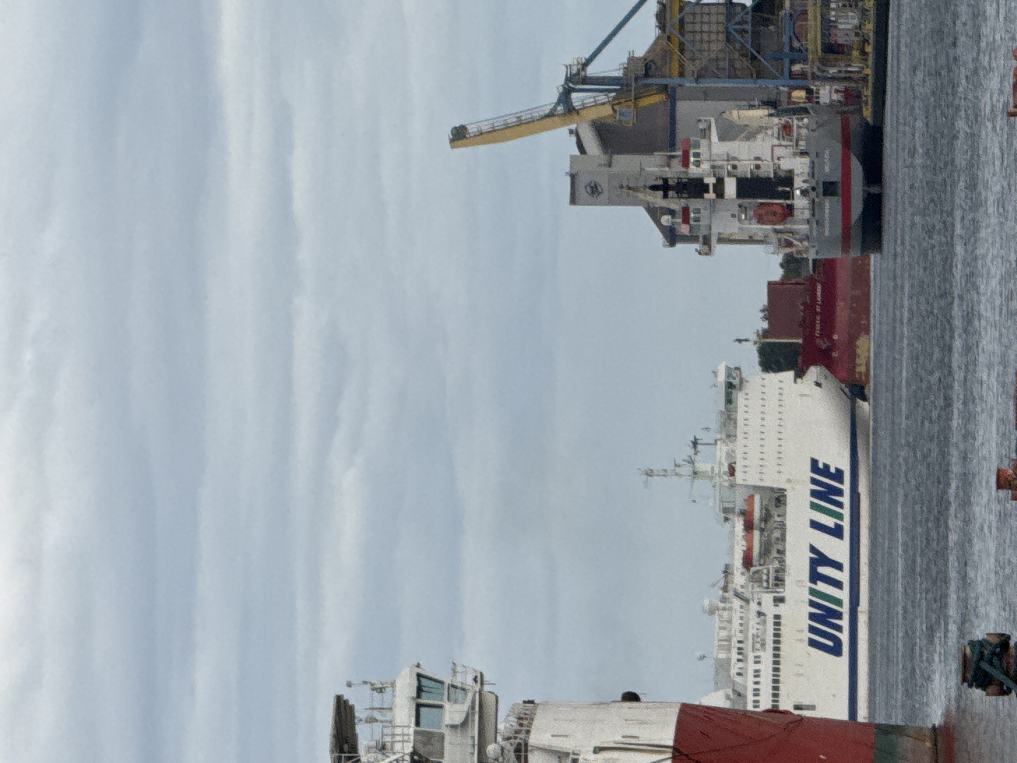 In Gdańsk, Poland, the image captures a maritime scene at port. A large white ferry with "UNITY LINE" emblazoned in blue and green letters is docked, its hull also featuring a blue stripe. To the left, the superstructure of another vessel, painted red and white, is visible. In the background, a port facility with industrial cranes, including a tall yellow one, stands against a cloudy sky. Smaller vessels and structures are also present in the water.
