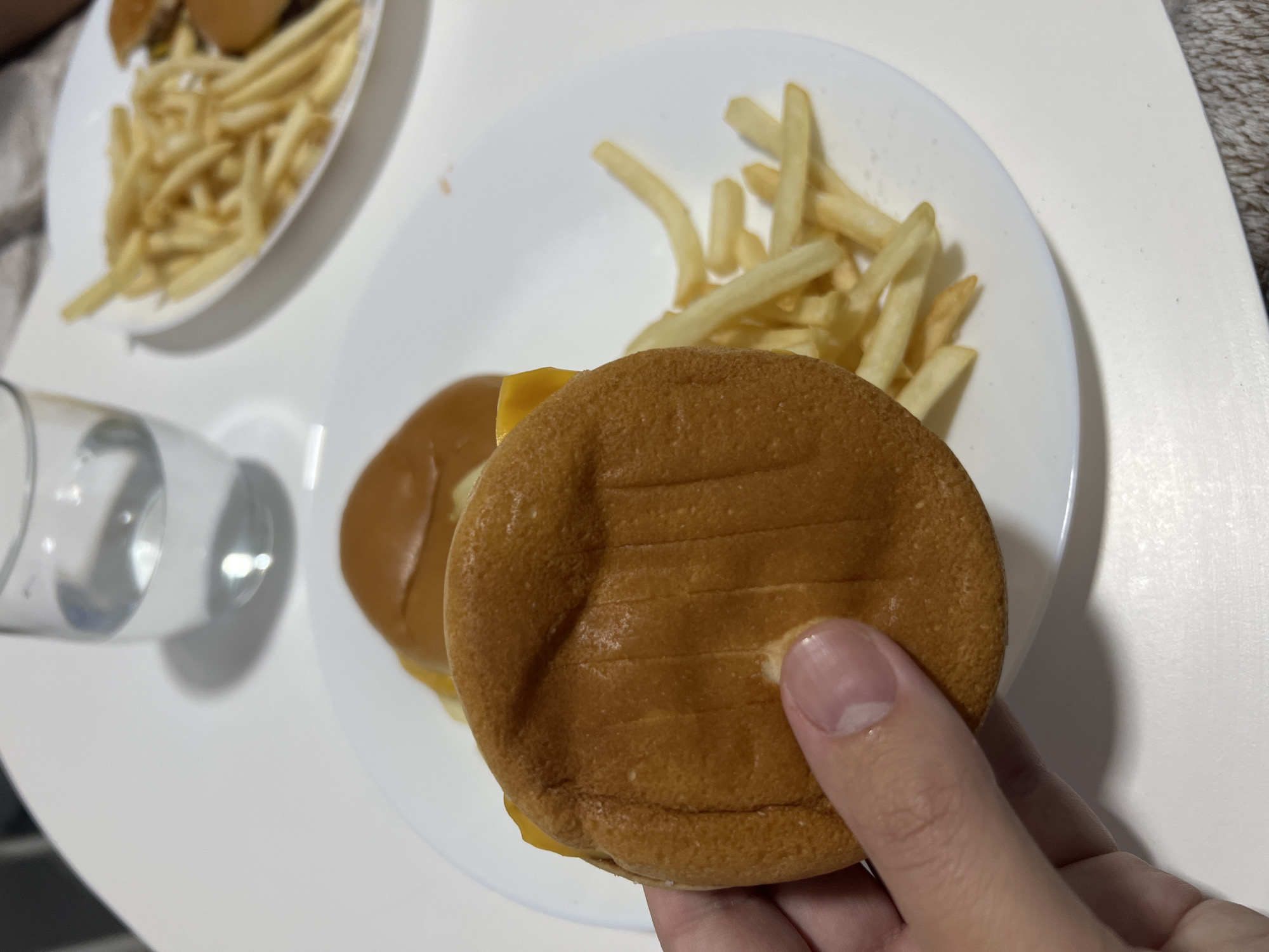 The image depicts a plate with a cheeseburger and french fries. A hand is holding the cheeseburger. Another plate with a cheeseburger and french fries is in the background, along with a glass of liquid. The setting appears to be a white table indoors. The location, Mogán, Spain, is not discernible from the visual information in the image.