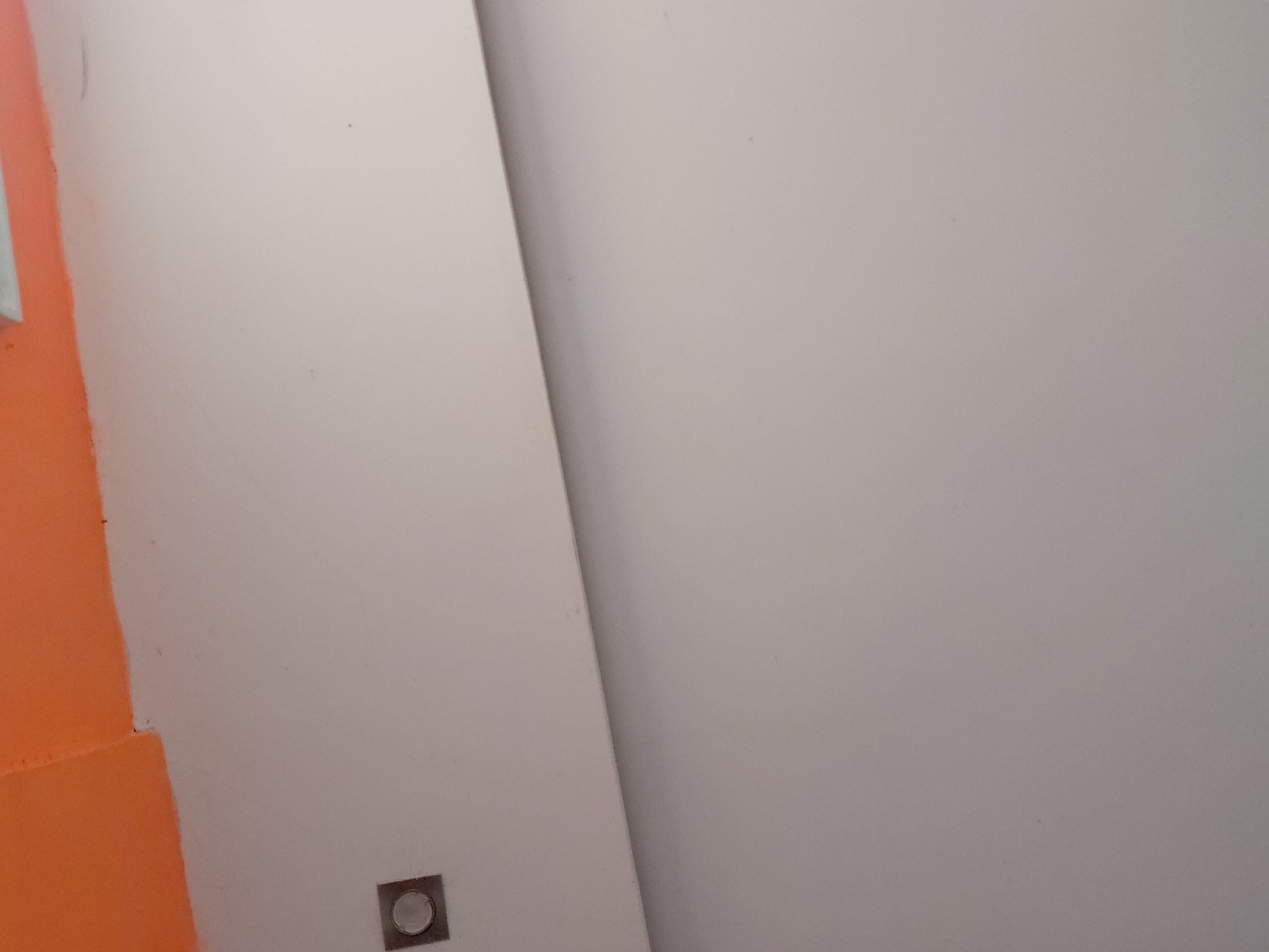 The image captures an abstract view of an interior architectural space, featuring distinct wall surfaces. On the left side, a vibrant orange wall section with visible brushstrokes and minor imperfections creates a bold contrast. This orange surface meets a much larger, off-white or light gray wall or ceiling plane, which occupies the majority of the frame. The boundary between these two light-colored surfaces is a sharp, dark diagonal line, suggesting a corner or a significant change in the room's geometry.
In the lower-left portion of the off-white wall, a small, square electrical fixture or outlet is visible. It features a dark or metallic frame with a circular, slightly reflective element at its center. The lighting is uniform, indicating either artificial illumination or diffuse natural light, and the time of day is not discernible. No people or activities are present in the static scene. The composition emphasizes lines, planes, and the contrast between the striking orange and the neutral background. No visible text is present. The location context provided is Santa Ana, Venezuela.