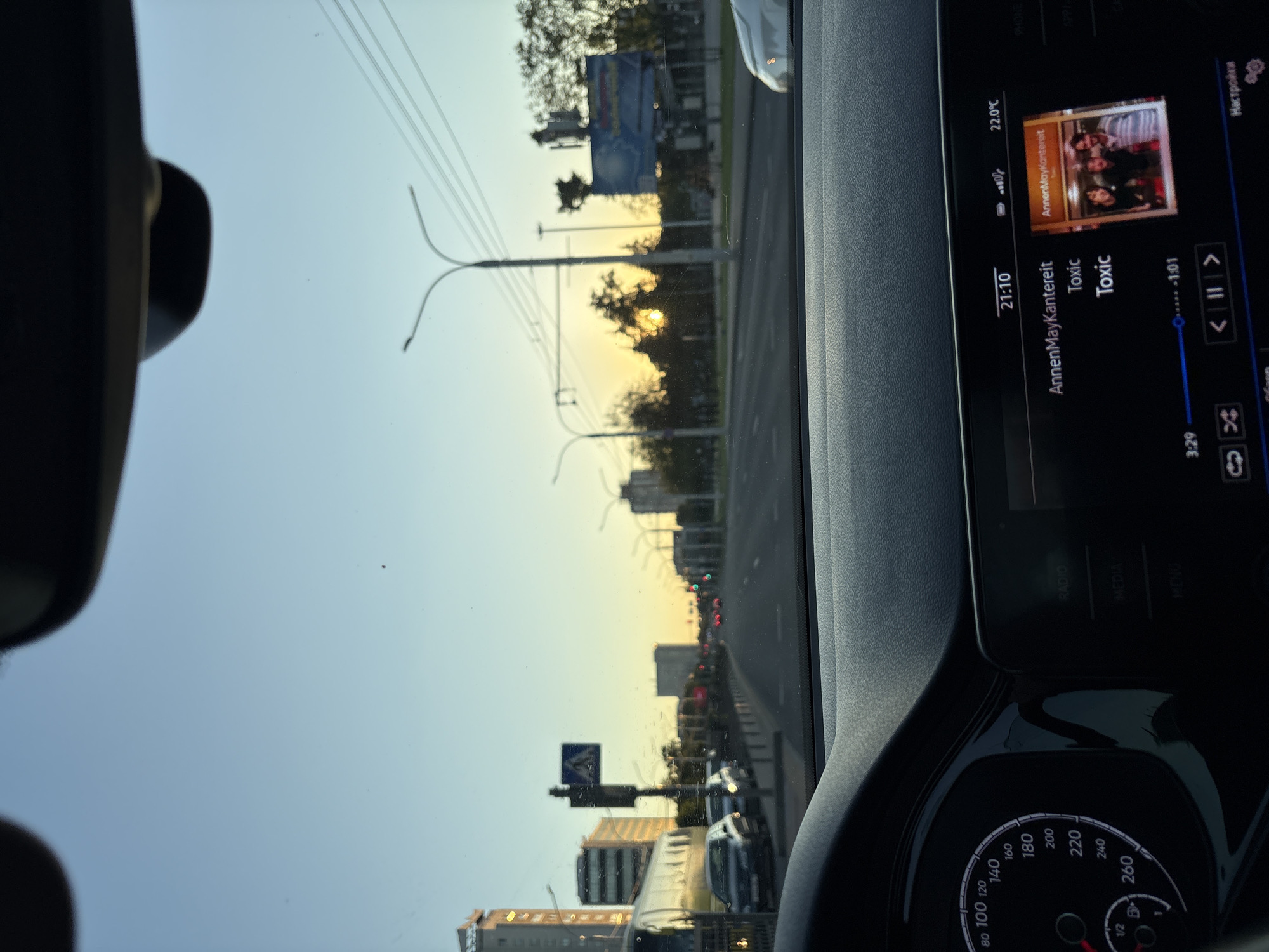 The image is taken from inside a car, looking out at a city street during what appears to be sunset. The dashboard is visible in the foreground, with a speedometer on the left and a central infotainment screen on the right. The infotainment screen displays the time as 21:10, a temperature of 22.0°C, and shows that a song by "AnnenMayKanterait" titled "Toxic" is playing, with playback at 3:29 out of 1:01. A small album art featuring three individuals is also displayed. The road ahead shows traffic and streetlights, with buildings and trees lining the street. The sky is a clear blue with a gradient towards yellow and orange near the horizon, indicative of sunset. The exterior view is slightly blurred, suggesting the car is in motion. The location is identified as Minsk, Belarus.