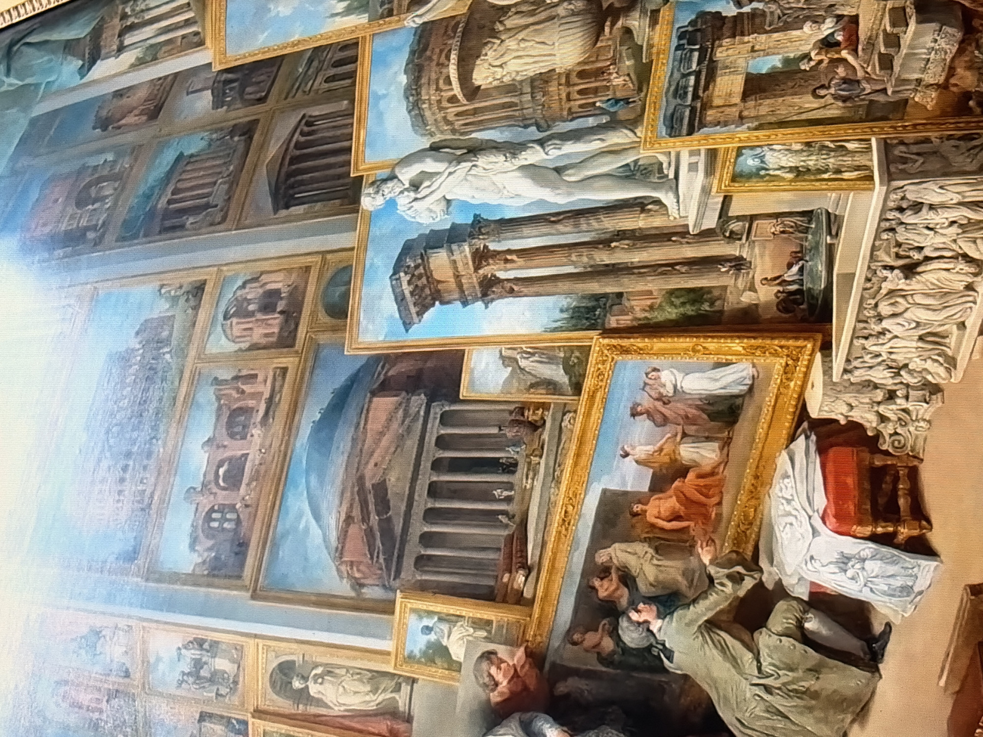 The painting depicts a gallery or exhibition of classical Roman architecture and sculptures. Several framed artworks are displayed on a wall and leaning against each other, showcasing various historical structures such as temples, arches, and amphitheatres, along with statues and reliefs. In the foreground, a man in historical attire is examining a framed painting, which displays figures in a classical setting. To his right, a red stool with a white cloth draped over it and a stack of framed artworks are present. The background is filled with more framed paintings, creating a dense display of Roman art and artifacts. The overall impression is a curated collection of historical imagery.