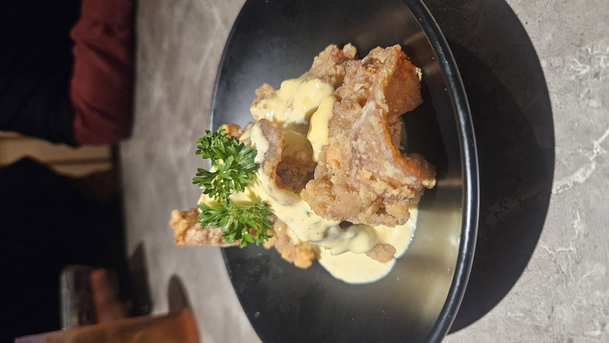 A close-up, eye-level shot focuses on a black, shallow bowl filled with golden-brown, crispy fried chicken pieces. A creamy, pale yellow sauce is generously drizzled over the chicken, and a sprig of fresh green parsley sits atop the dish as a garnish. The bowl rests on a textured, grey surface that appears to be a table with a marble-like pattern. In the blurred background, the dark clothing of two individuals is visible, suggesting a dimly lit indoor setting, possibly a restaurant. The lighting casts a prominent shadow from the bowl onto the table in the foreground. The scene does not provide any specific information to determine the location as Bekasi, Indonesia.