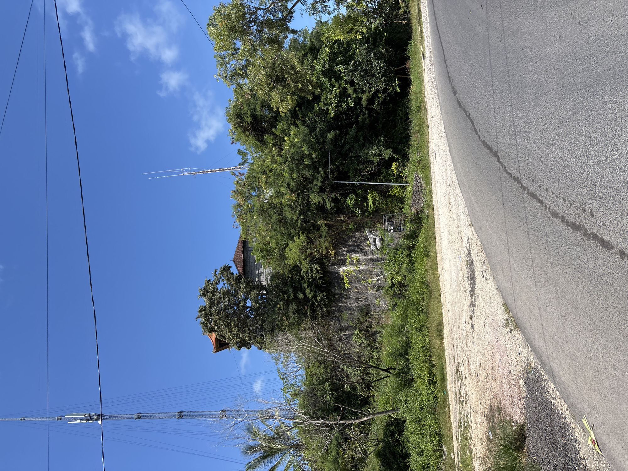 The image shows a roadside scene in Kuta Selatan, Indonesia. A paved road runs along the right edge of the frame. Beside the road is a gravel shoulder with vegetation. Beyond the shoulder, a steep, vegetated slope leads up to a structure with a red-tiled roof, partially obscured by trees. Power lines are visible in the foreground and antennas extend into the blue sky.