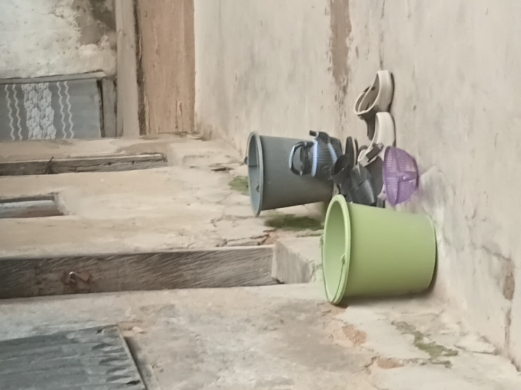 The image depicts an exterior scene in Jalingo, Nigeria, focusing on a section of a building and its immediate surroundings. To the left, a rough concrete wall is visible, textured with patches of peeling paint and moss. A corrugated metal sheet is fixed to this wall. Adjacent to the wall is a light green plastic bucket. Further to the right, a wooden door is set into the building, secured with a padlock. Next to the door, and slightly in front of it, are two large plastic buckets, one gray and one darker gray, with a black and white striped pitcher or kettle placed in front of the darker gray bucket. A pair of dark shoes is positioned next to the pitcher. In the foreground, to the right of the green bucket, is a small, translucent purple plastic bowl. Further to the right, two pairs of shoes are visible: one pair of white slip-on shoes and another pair of dark clogs. The ground is a concrete surface, showing some damp patches. The background reveals more of the building with a closed doorway and a patterned curtain or covering.