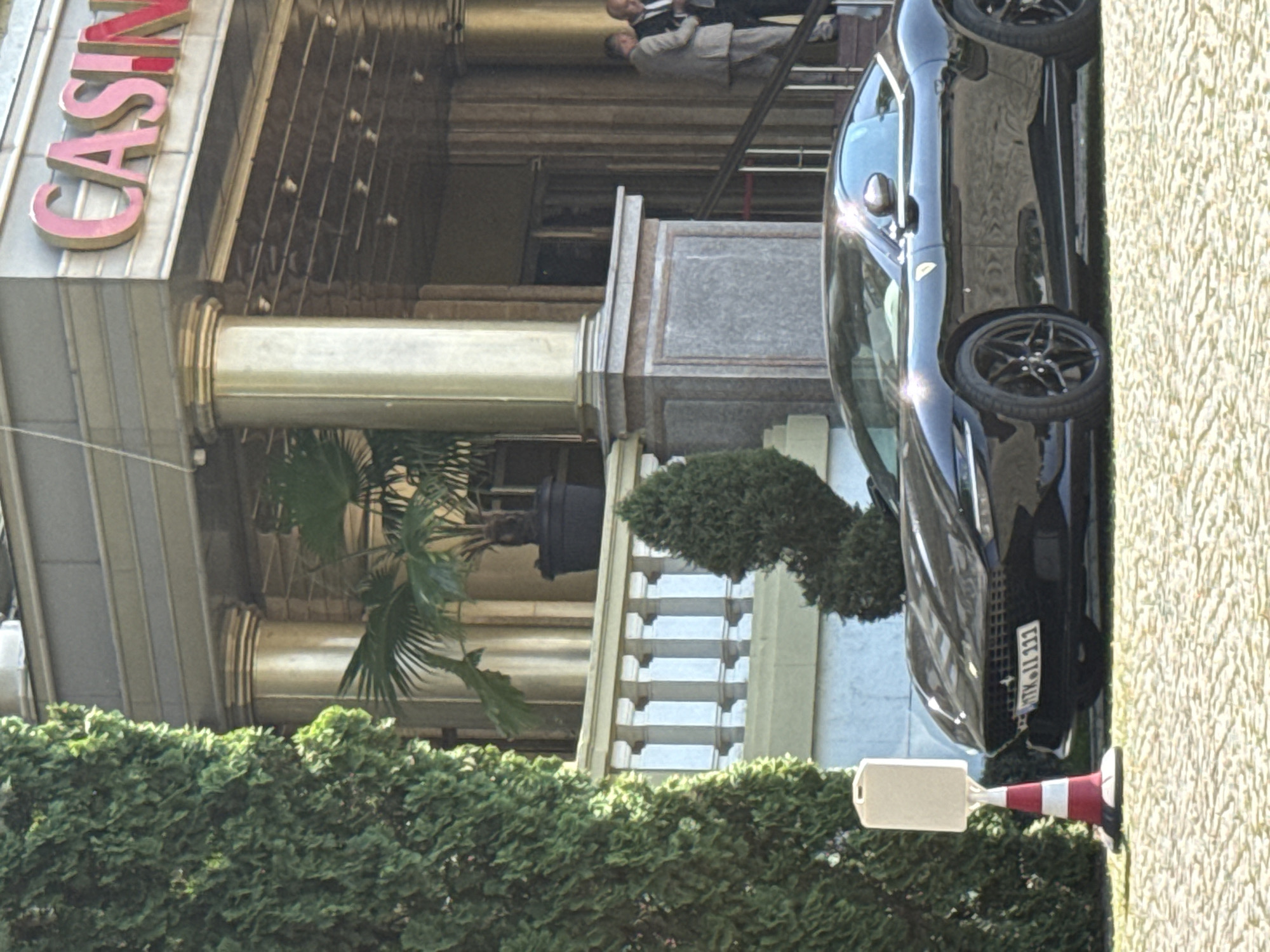 A color photograph shows the Casino International Plovdiv in Bulgaria, featuring the word "Casino" in large, pink, three-dimensional letters on the building's facade. The building has a classical architectural style with gold-colored columns. A dark-colored car, potentially black or a very dark shade of gray, is parked to the right of the building. A section of a green hedge or bush is visible in the lower-left foreground. In the background, two people are standing on the casino steps. A traffic cone, with a white sign attached to it, is next to the car.