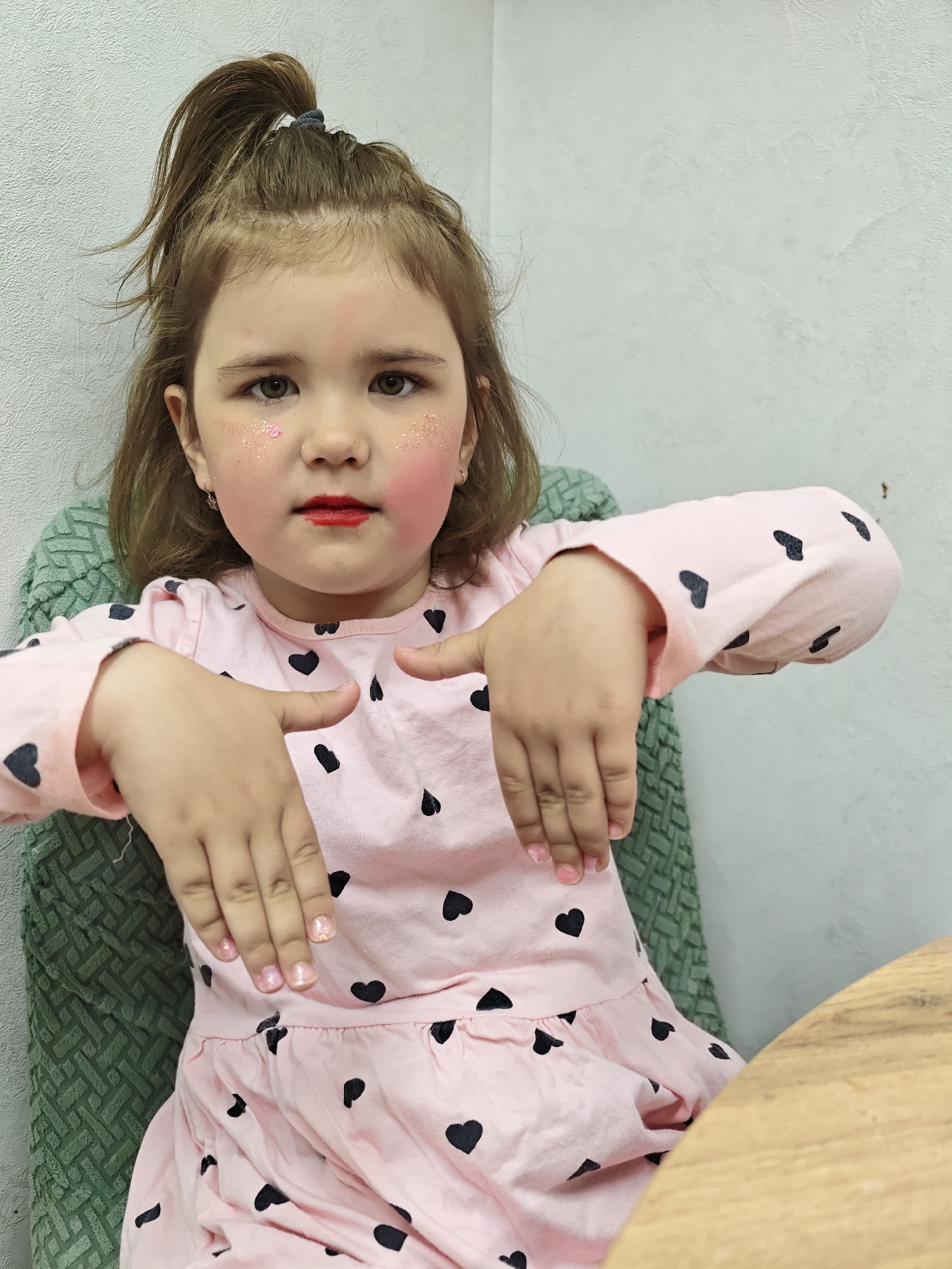 A young girl with brown hair styled in a high ponytail sits in a green textured chair at a wooden table. She wears a pink long-sleeved dress patterned with black hearts. The girl has pink blush on her cheeks and glitter on her face near her eyes, and red lipstick is smeared on her lips. Her fingernails are painted with a glittery pink polish. The background consists of a light gray wall. This photo was taken in Chaikovsky, Russia.