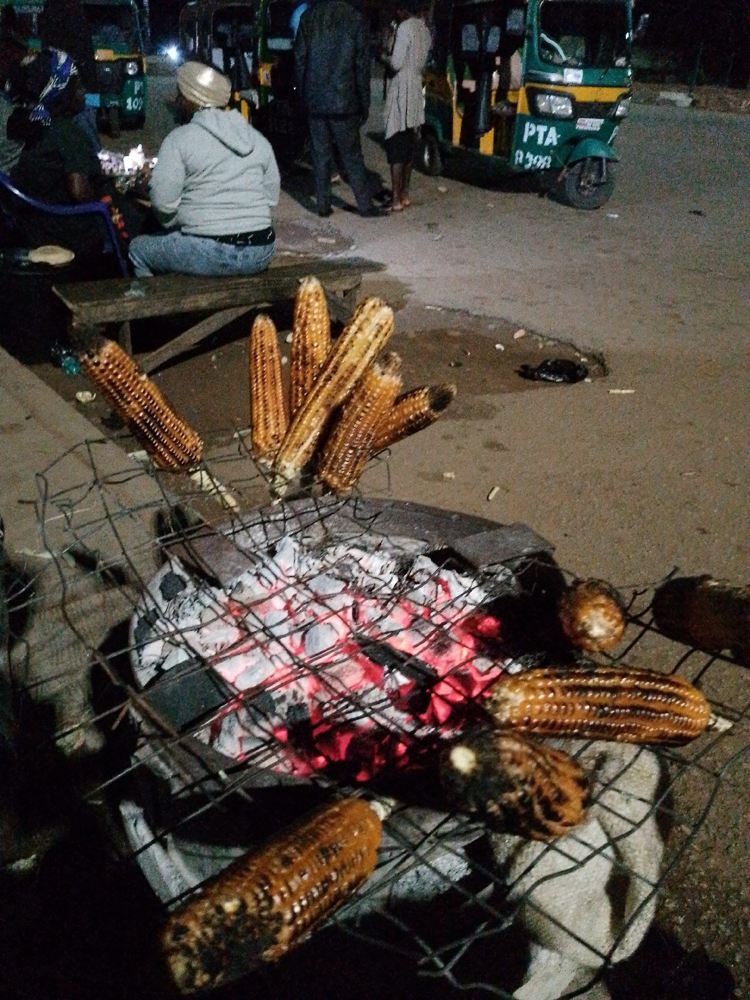 The image depicts an outdoor scene at night, likely in Bukuru, Nigeria, where corn is being grilled over hot coals. The main subjects are several ears of corn arranged on a wire grill, with glowing red embers visible beneath. The corn appears to be roasted, with some kernels showing char marks.
In the background, three individuals are present. One person, wearing a grey hooded sweatshirt and jeans, sits on a wooden bench. Another individual, partially visible, is dressed in dark clothing and is standing. A third person, wearing a light-colored garment, is also standing. The individuals appear to be interacting or observing the grilling process.
Several vehicles are also visible, including a green and yellow auto-rickshaw with the text "PTA 1202" on its side. Another auto-rickshaw is partially visible to the left. The setting appears to be a street or roadside area, characterized by the asphalt ground and the presence of vehicles and people. The time of day is clearly night, indicated by the artificial lighting and the dark sky. The overall mood seems to be casual, possibly a street food vendor preparing and serving grilled corn to customers or passersby.