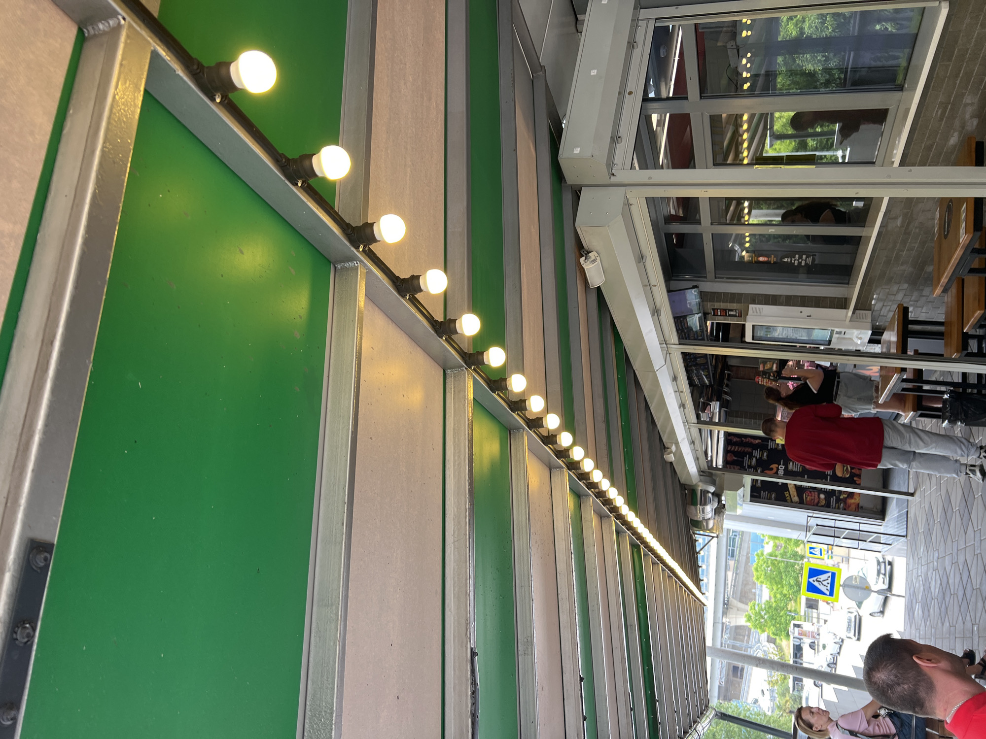The image depicts an outdoor seating area under a green awning supported by metal beams. A string of round light bulbs is illuminated along one of the beams, casting a warm glow. In the background, several people are visible. A woman in a pink shirt is seated on the left, facing away from the camera. A man in a red shirt and gray pants stands in front of a glass-enclosed food service counter on the right. Two other individuals are visible through the glass. Outside the seating area, a street scene is visible with cars, a bus, and pedestrian crossing signs. This scene is consistent with an outdoor dining establishment in Sochi, Russia, possibly near a park or recreational area.
