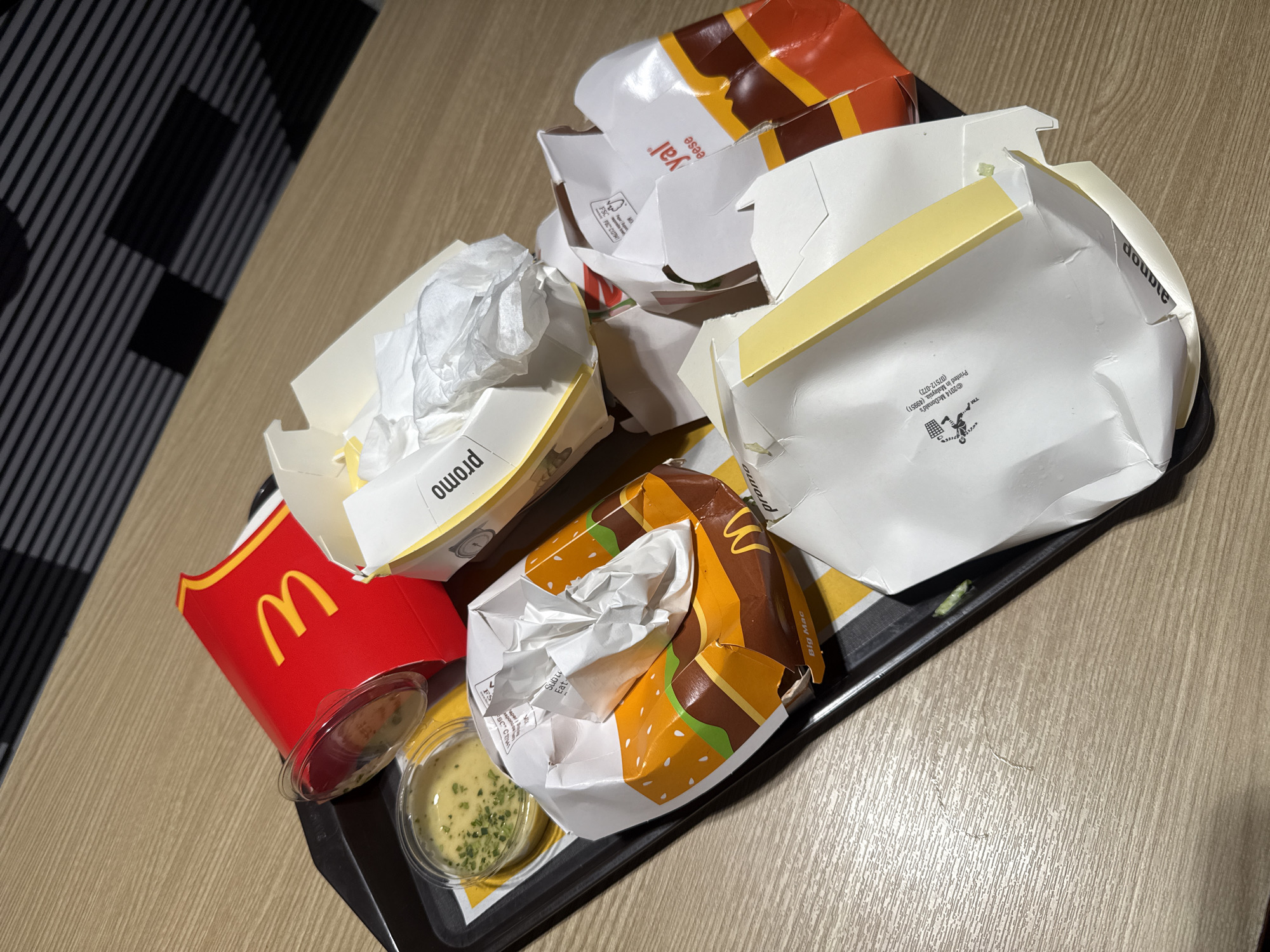The image shows a used McDonald's tray with several empty food containers and a small cup of dipping sauce on a wooden table. The containers include a red fries box with the McDonald's logo, a crumpled Big Mac box, and several other white paper boxes, some with text like "Royal" and "Double". The dipping sauce appears to be a creamy yellow sauce with green herbs. The background shows a patterned wall and a striped ceiling. The scene is consistent with a fast-food dining area, possibly at a McDonald's restaurant. The specific location in Da Lat, Vietnam is not discernible from the image itself.