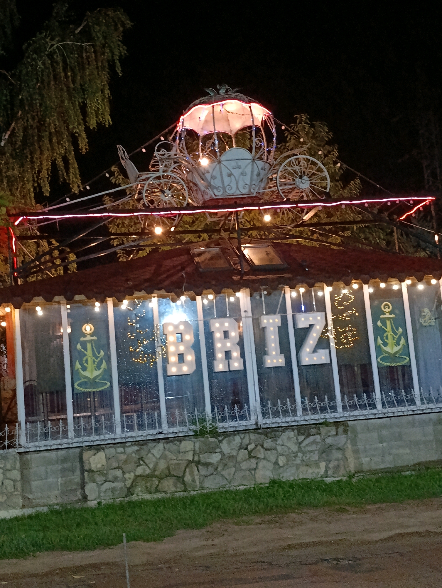 The image depicts a nighttime scene of a building exterior, likely a cafe or restaurant, in Korzhevo, Moldova. Above the building's roof, a decorative white carriage illuminated by lights is visible. The building itself has a dark roof and large windows, through which interior lights and decorations can be seen. Prominently displayed on the facade are illuminated white letters spelling "BRIZ". On either side of the letters, the windows feature green anchor motifs. The building is constructed on a stone foundation, with a grassy area in front and a dirt path or road. The overall impression is of an outdoor establishment decorated for evening ambiance.