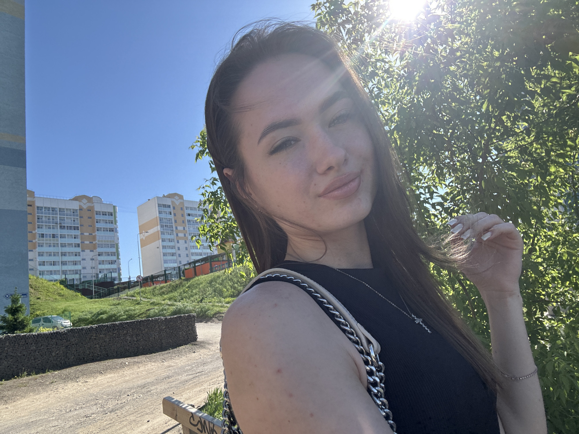 A bright outdoor selfie features a young woman with long, dark brown hair, fair skin, and light makeup, including defined eyebrows and pinkish lip color. She is looking directly at the camera with a soft, closed-mouth smile. She wears a black sleeveless top, a silver cross pendant necklace, and carries a silver chain shoulder bag. Her right hand, adorned with a delicate silver bracelet and long, light-colored fingernails, is casually raised near her hair on the right side of the frame. Her shoulder and arm are visible, showing a few small blemishes.
The setting is an urban or suburban area under a clear, bright blue sky. The sun is strongly present on the right side of the image, creating significant lens flare and backlighting the woman and the trees behind her. In the background, modern multi-story apartment buildings with light-colored facades are visible. Green grassy slopes and trees line a dirt or gravel path. A prominent gabion wall, constructed from wire cages filled with rocks, runs horizontally across the middle-ground. Parts of a parked white car and streetlights are also visible. A partially obscured wooden bench with some illegible carved or painted text is present in the lower right corner. The bright sunlight suggests it is daytime.