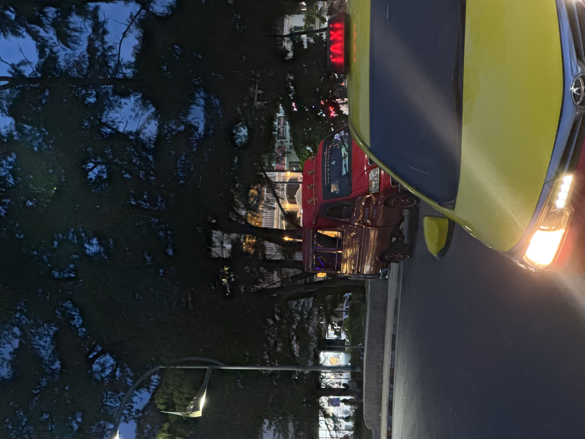 In Karon, Thailand, a yellow taxi is parked on an asphalt road at dusk. The taxi's headlights are illuminated, and its "TAXI" sign is lit in red. A red pickup truck, possibly a tuk-tuk, is parked behind the yellow taxi, with a sign on its windshield containing Thai script. Trees with dark green foliage are visible in the background, obscuring a row of buildings illuminated by artificial lights. A street lamp with a white light is also present. The sky is a deep blue, indicative of twilight. The overall scene suggests a street in a busy area with ongoing traffic.
