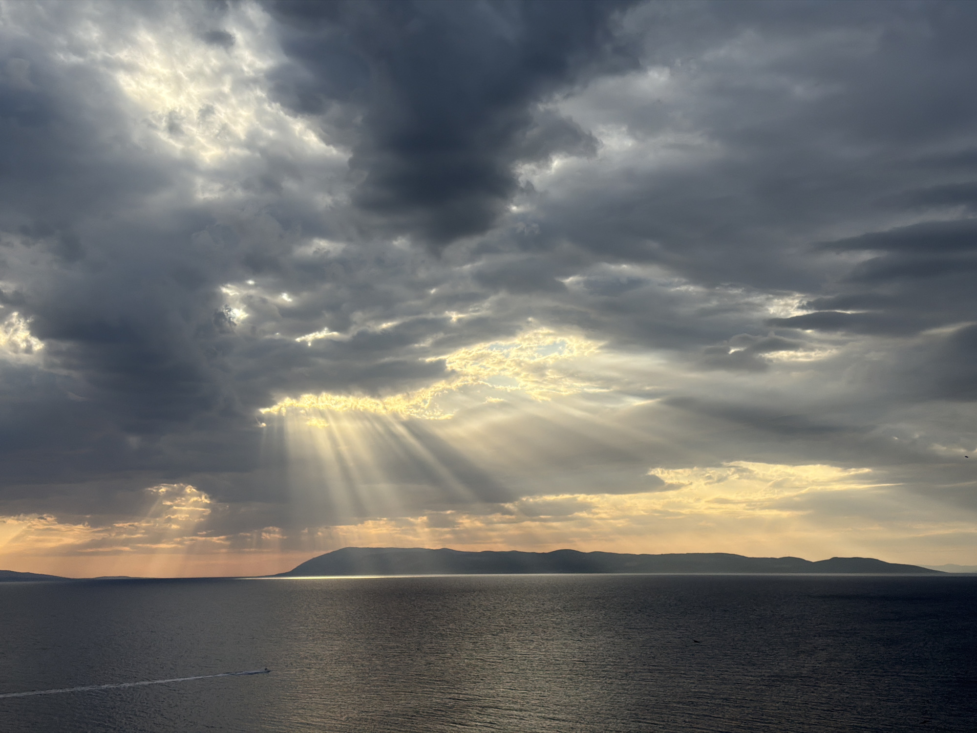 The image captures a dramatic sunset over the Adriatic Sea, viewed from Općina Podgora, Croatia. Dark, brooding clouds dominate the sky, but shafts of sunlight pierce through them, creating a powerful crepuscular effect. These sunbeams illuminate the sea and cast long shadows on the water's surface. In the distance, a long, dark silhouette of mountains or islands stretches across the horizon. The water itself is a deep, dark blue, with a single white wake from a small boat visible on the left side of the frame. Two birds are also visible in flight against the sky. The overall impression is one of both foreboding and awe-inspiring natural beauty.