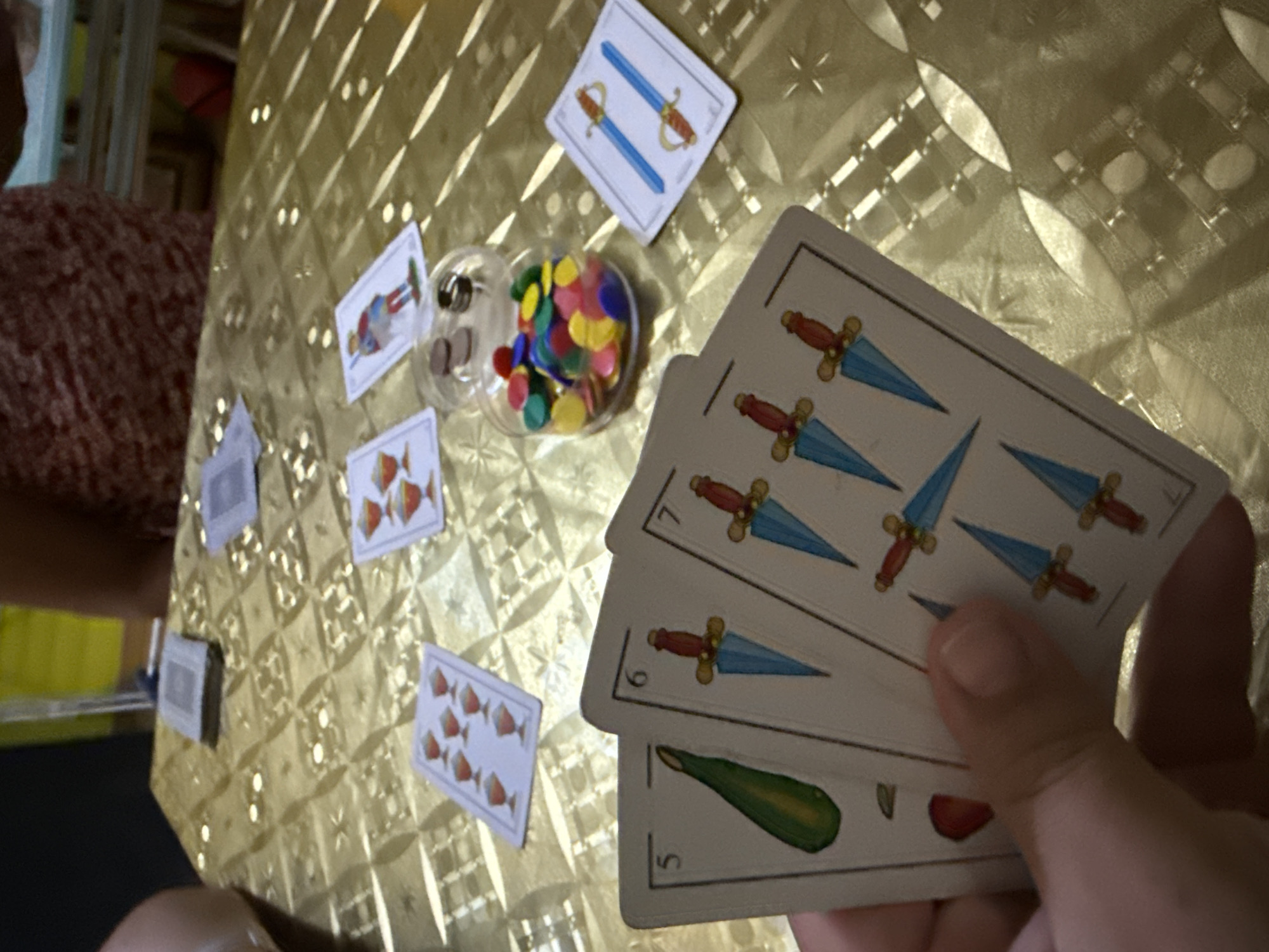 A close-up, high-angle, indoor shot captures a table covered with a gold, textured cloth. The table displays several playing cards, some face up and some face down. In the foreground, a hand is holding three cards from a Spanish deck: a '5' card with a green vegetable (likely a bean pod), a '6' card with three swords, and a '7' card with four swords. Scattered across the table are additional cards, including a '7' of cups, a '4' of cups, a 'page' of clubs, and a '2' of swords. A small glass jar filled with colorful game tokens (red, yellow, blue, and green) sits near the cards, with a few tokens and a ring resting on top. The background is blurred, showing parts of people and furniture. The scene is likely a domestic setting, possibly a game night or casual gathering, in Getafe, Spain, though there are no specific visual cues to confirm the exact location within the city.