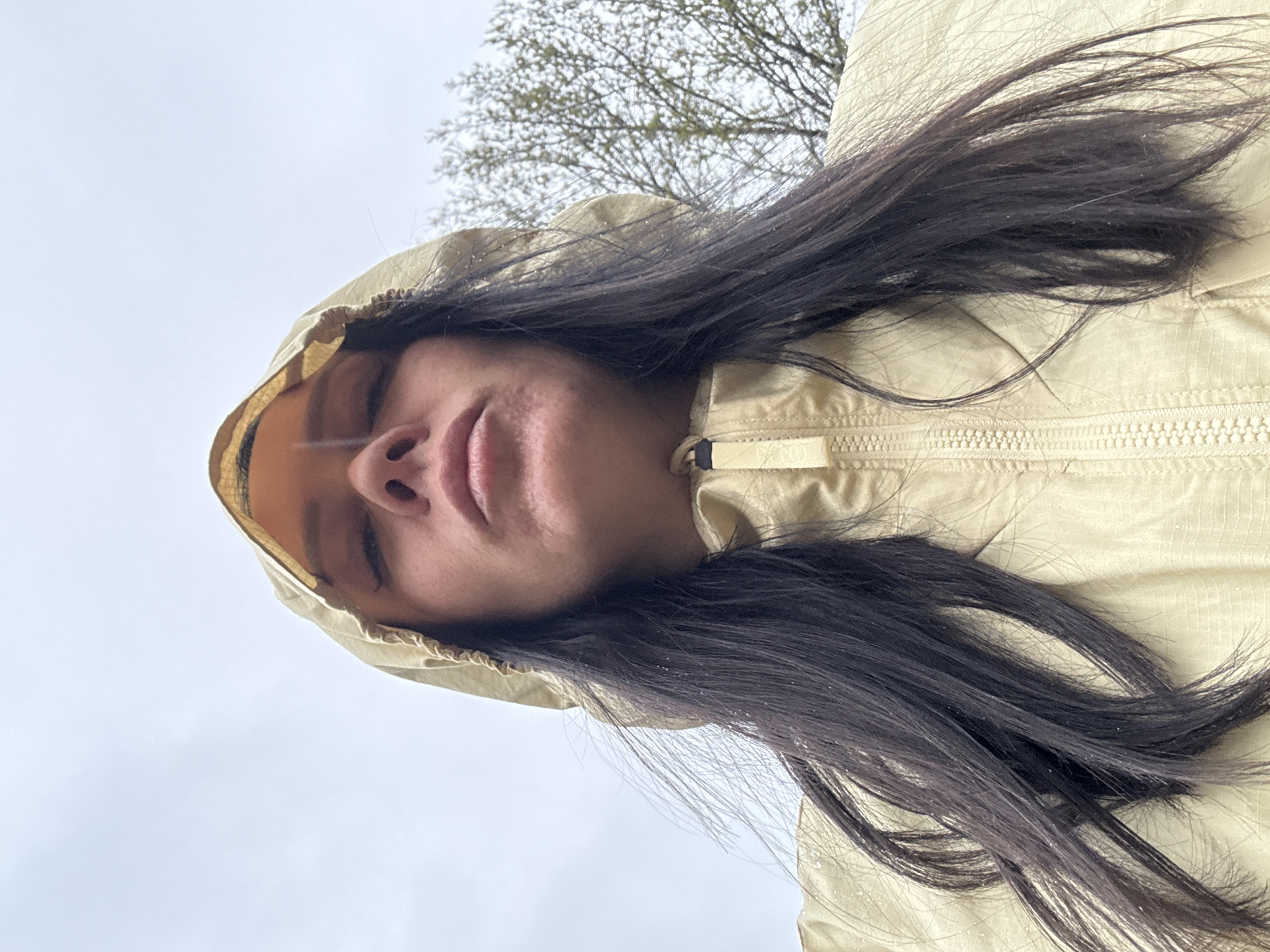 A woman with long dark hair and fair skin is centered in the frame. She is wearing a hooded jacket in a light beige color, with the hood up. Her eyes are closed. Above her head, a bare tree branch is visible against a gray sky. The image is oriented at an angle, appearing as though the woman is looking directly downward toward the camera. It is not possible to discern the location in Chaikovsky, Russia, or any actions or interactions from this single image.