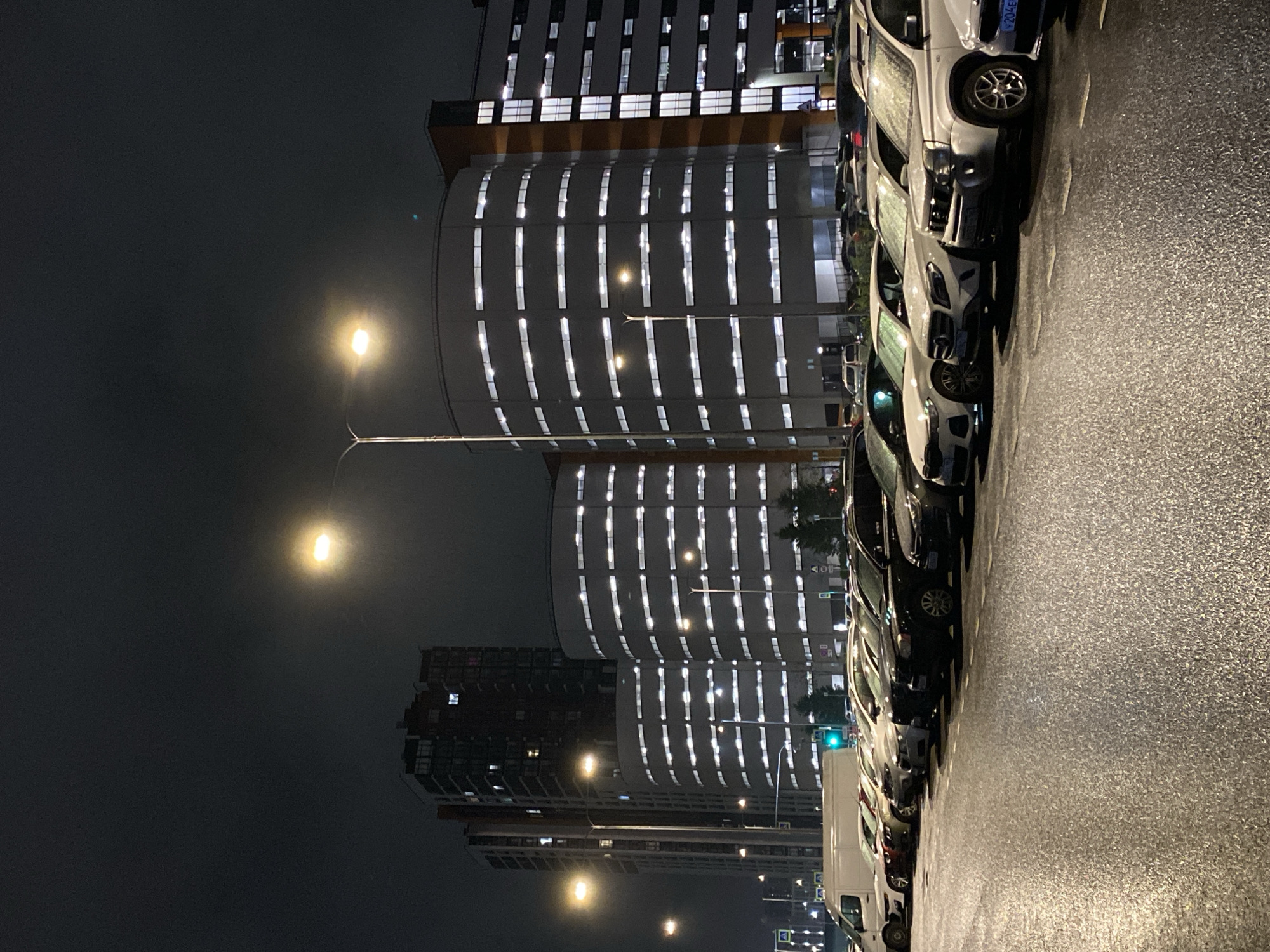 The image depicts a nighttime urban scene, likely taken in Saint Petersburg, Russia. The foreground features a wet asphalt surface, reflecting the surrounding lights. A row of parked cars lines the right side of the image. Behind the cars are several buildings, including a taller structure with a distinct rounded facade and numerous vertical rows of illuminated windows. Streetlights are visible, casting a glow on the buildings and the surrounding area. The sky is dark and overcast, contributing to the overall dim and wet atmosphere.
