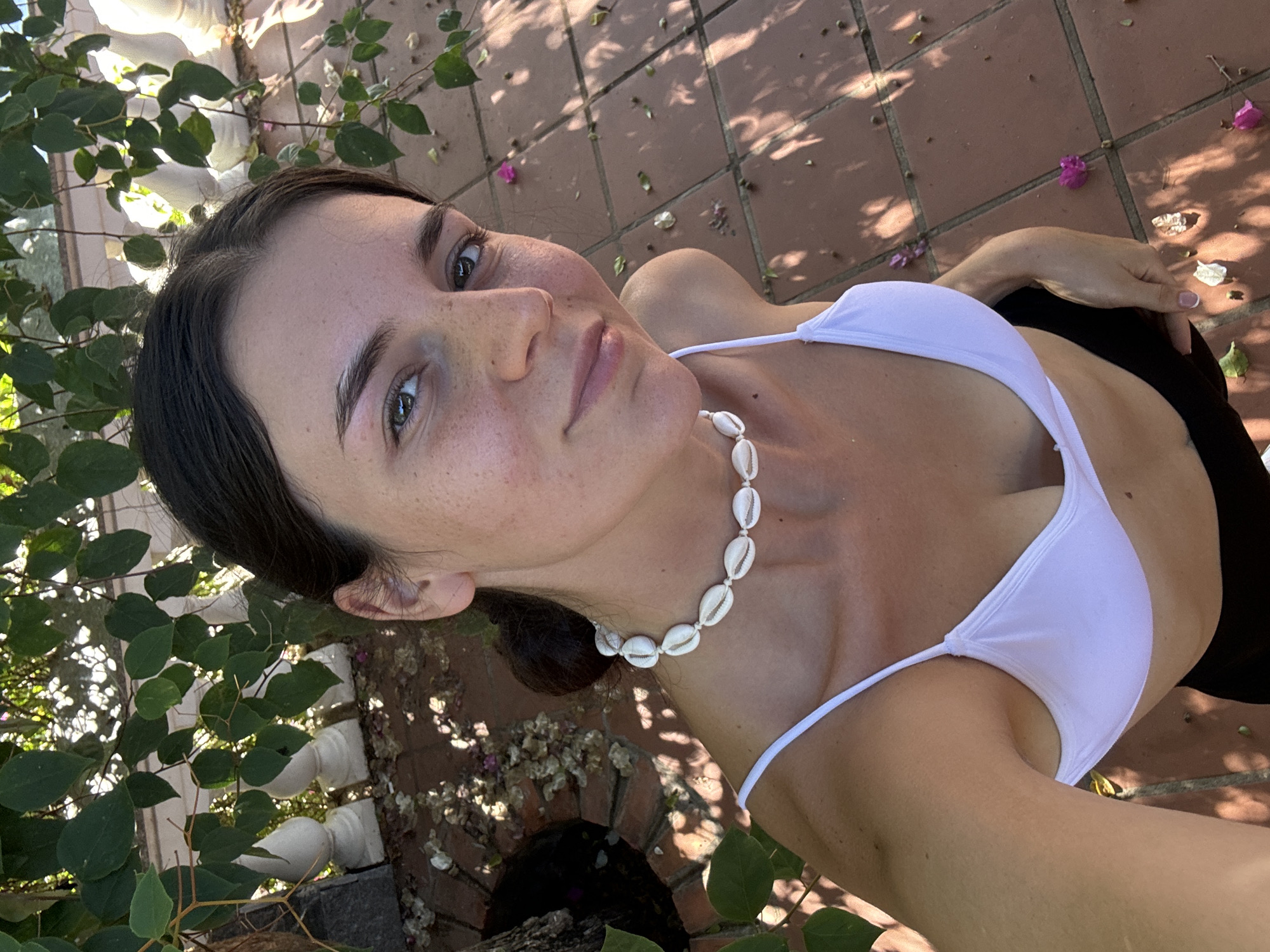A woman is posing for a selfie. She has dark hair pulled back from her face, freckles, and a seashell necklace. She is wearing a white bikini top and black bottoms. The setting appears to be an outdoor area with brick flooring and a garden with plants. The location may be Mũi Né, Vietnam.