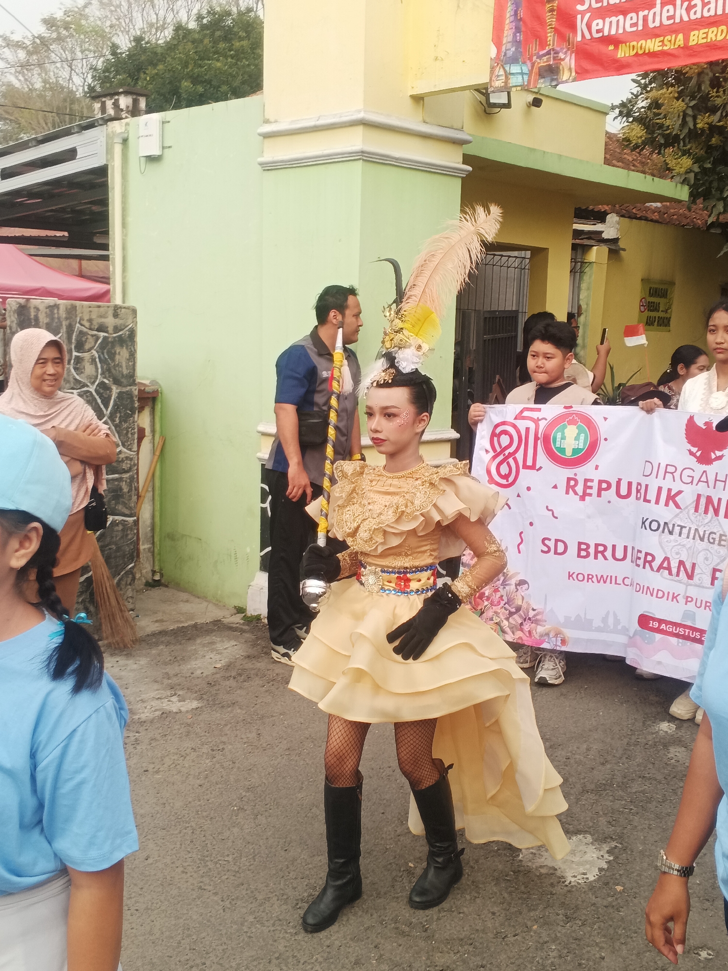 In Sokanegara, Indonesia, a girl in a gold, ruffled dress and black boots stands holding a baton. She has on black gloves and fishnet stockings. Behind her, a banner reads "DIRGAH UT REPUBLIK INDONESIA" and other text. A young boy holds the banner up. A woman in a headscarf stands to the left of the girl, and a man in a blue shirt is behind her. In the background, a building with a yellow and light green facade is visible. The ground is paved and appears to be a street or pathway.